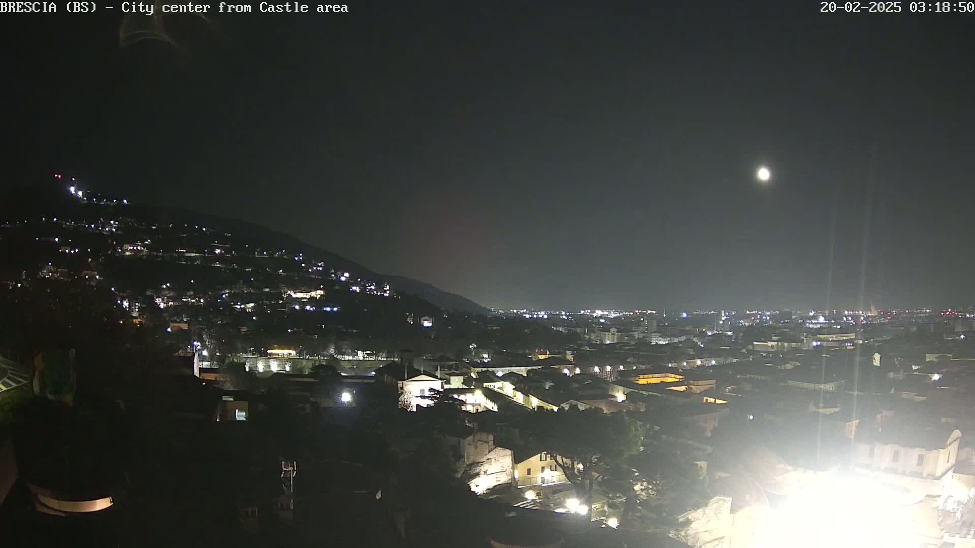A nighttime view of a city nestled against a hillside, illuminated by streetlights under a clear sky with a visible moon.