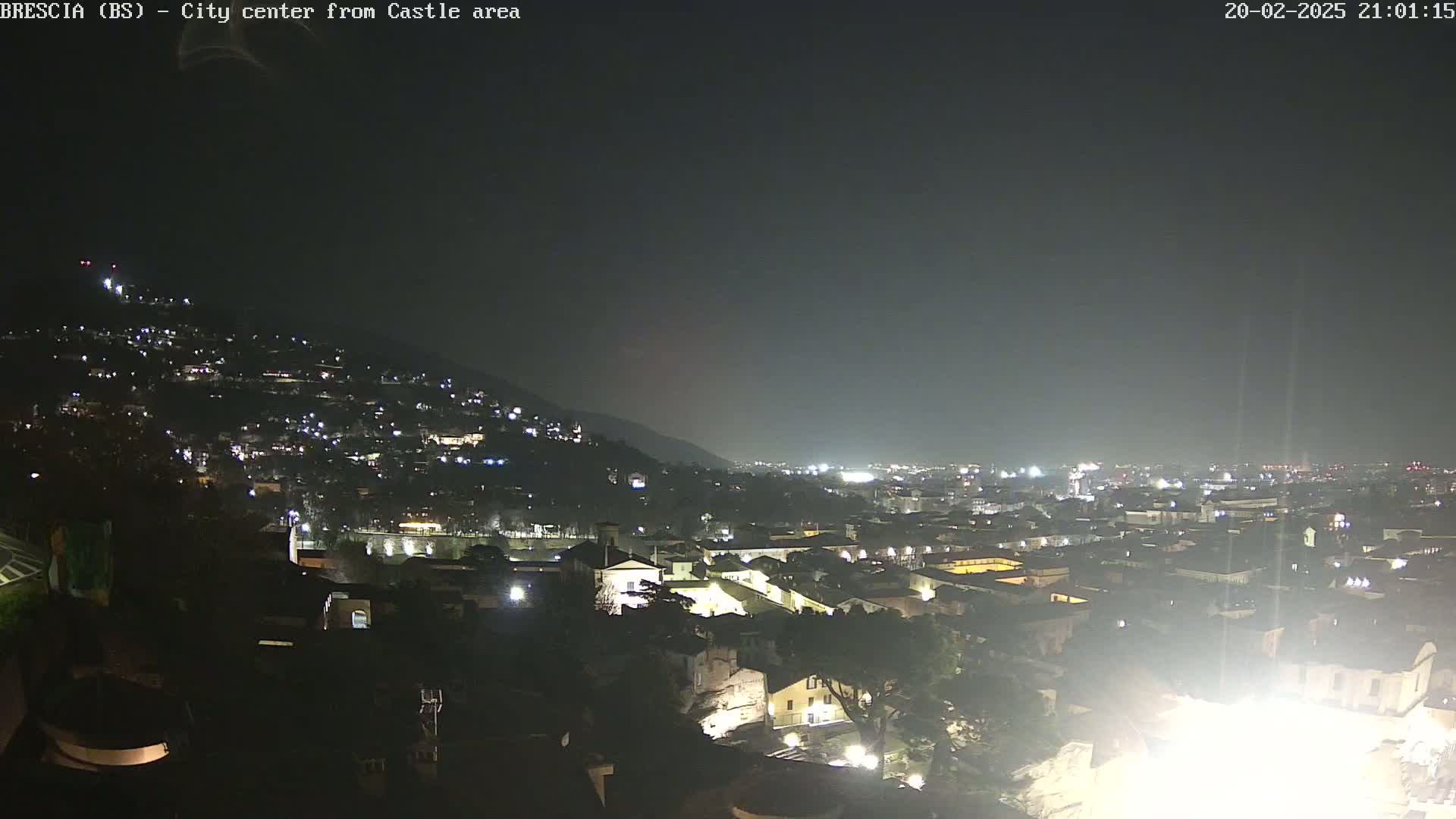 A nighttime view of a city illuminated by lights, nestled against a dark hill under a cloudy sky.