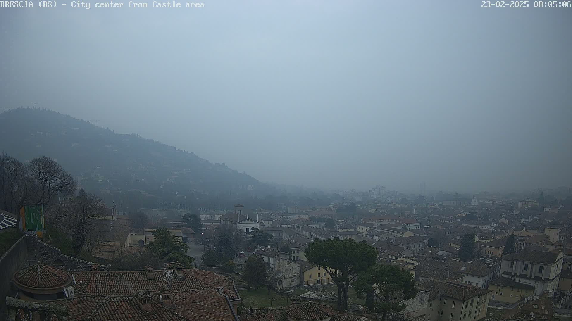 A hazy, overcast view shows a city nestled in a valley below a hill, with many brown-tiled roofs visible.