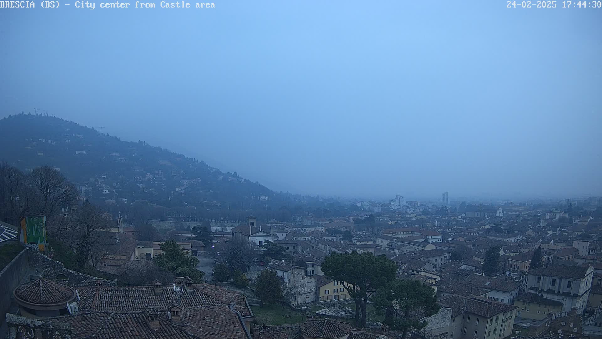A hazy, bluish-grey city sprawls across a hillside, viewed from a higher vantage point, with a hill in the background.