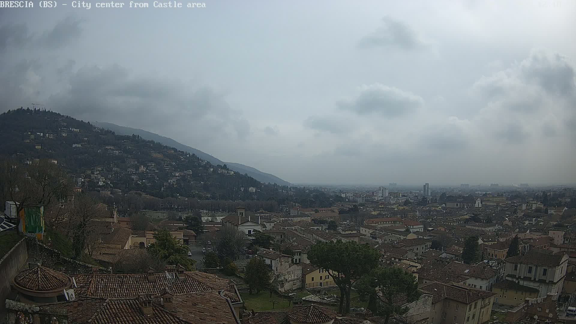 A sprawling city with red-tiled roofs nestled beneath a hill, viewed on an overcast day.