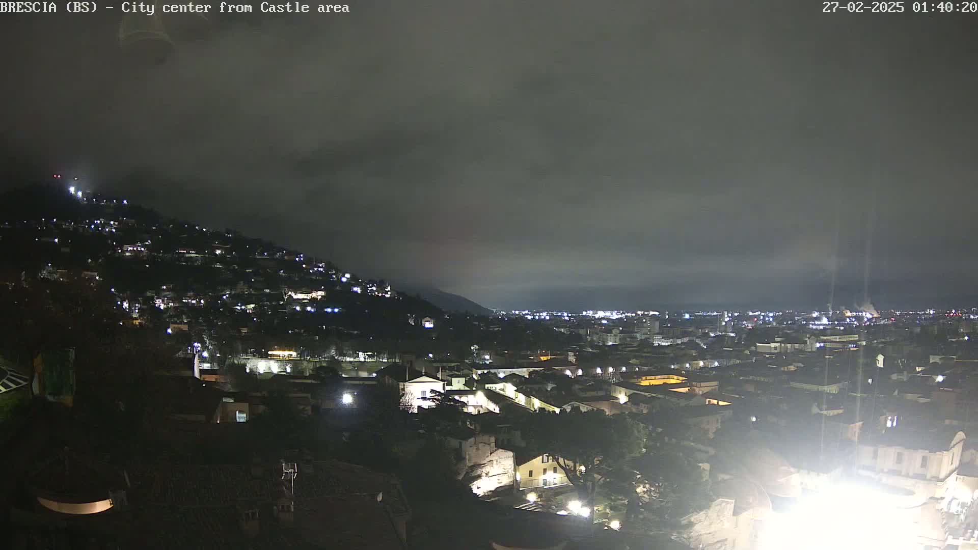 A nighttime view of a city nestled on a hillside under a cloudy sky, with numerous lights illuminating the buildings and streets below.