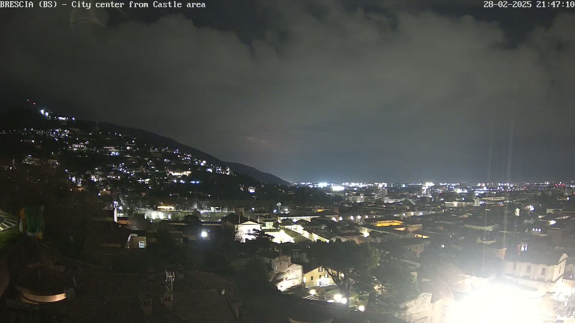 A nighttime view of a city nestled against a hill, under a mostly cloudy sky.