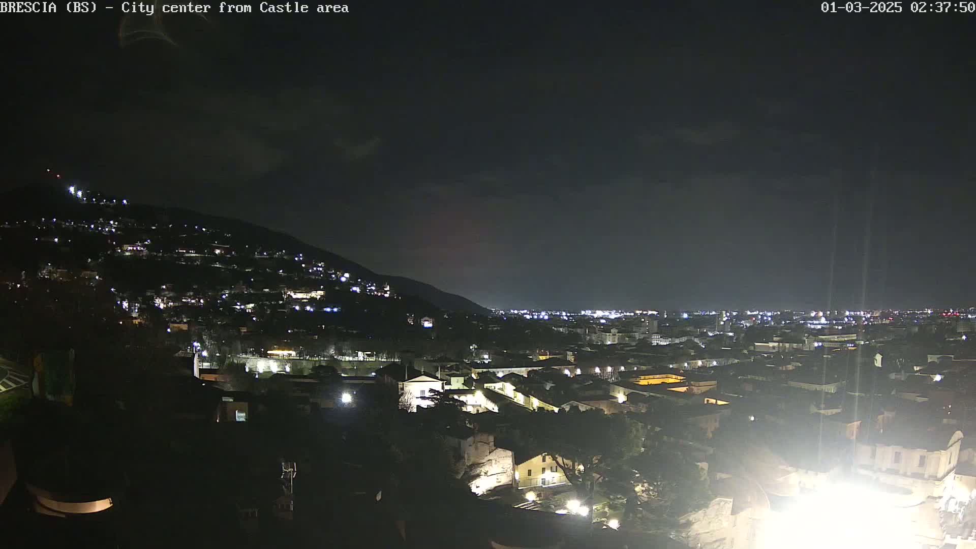 A nighttime, long shot of a city nestled against a hill, illuminated by scattered lights under a dark sky.