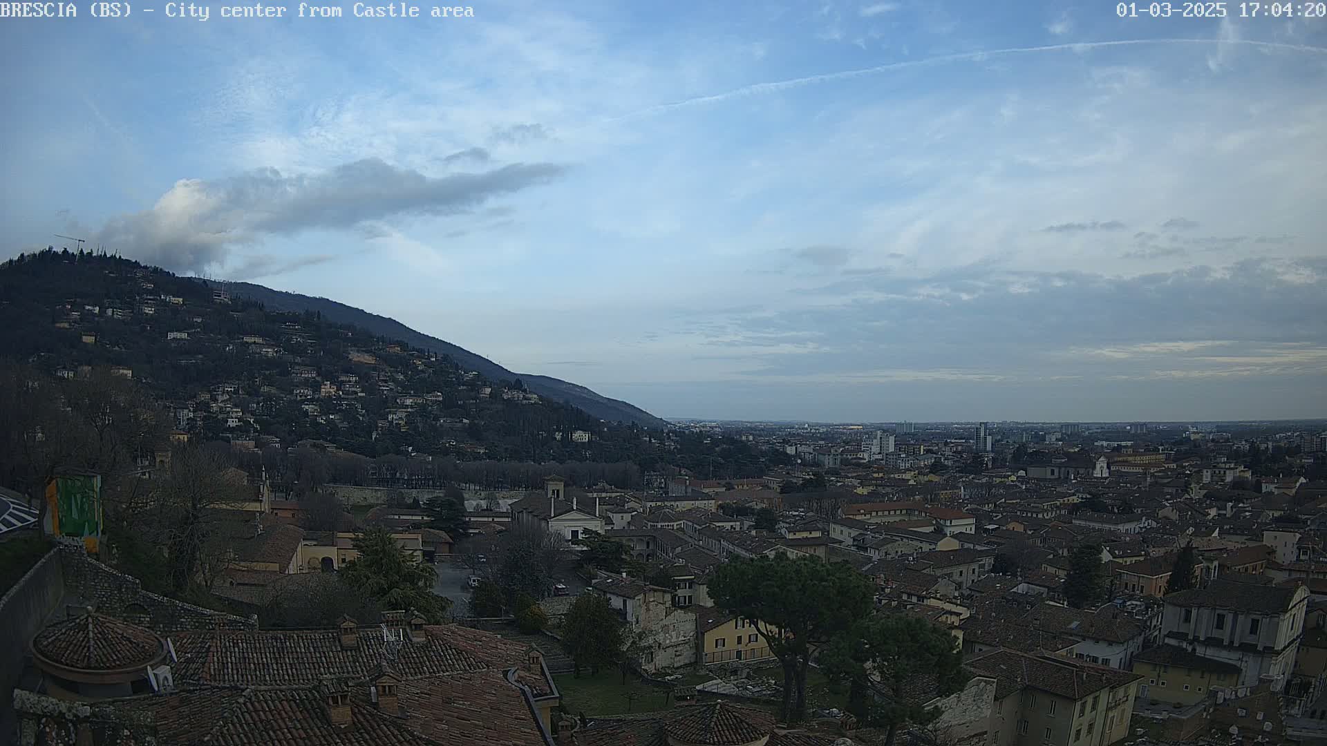 A sprawling city with terracotta rooftops nestles beneath a hill dotted with houses, all under a mostly clear, pale blue sky.