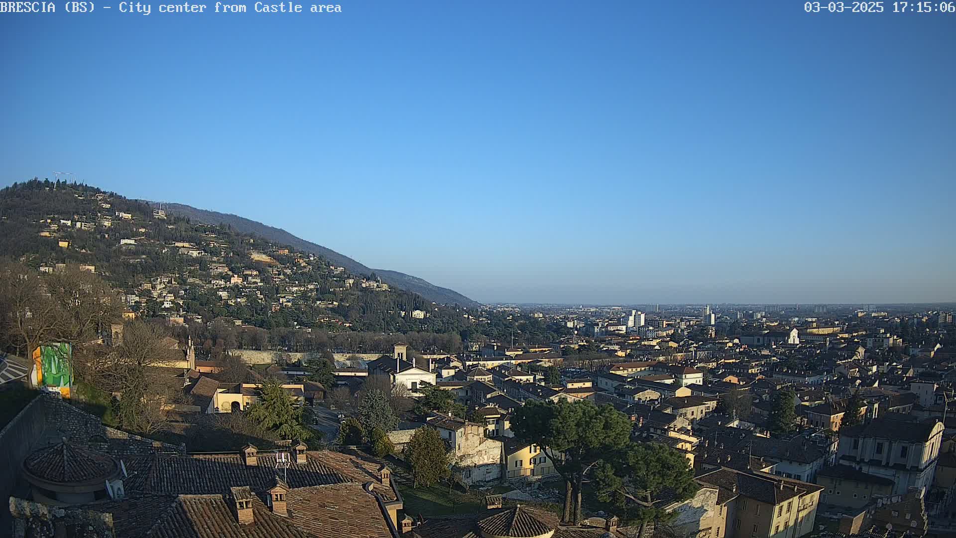 A sprawling city nestled in a valley beneath a clear blue sky and a hillside dotted with buildings, viewed from a high vantage point.