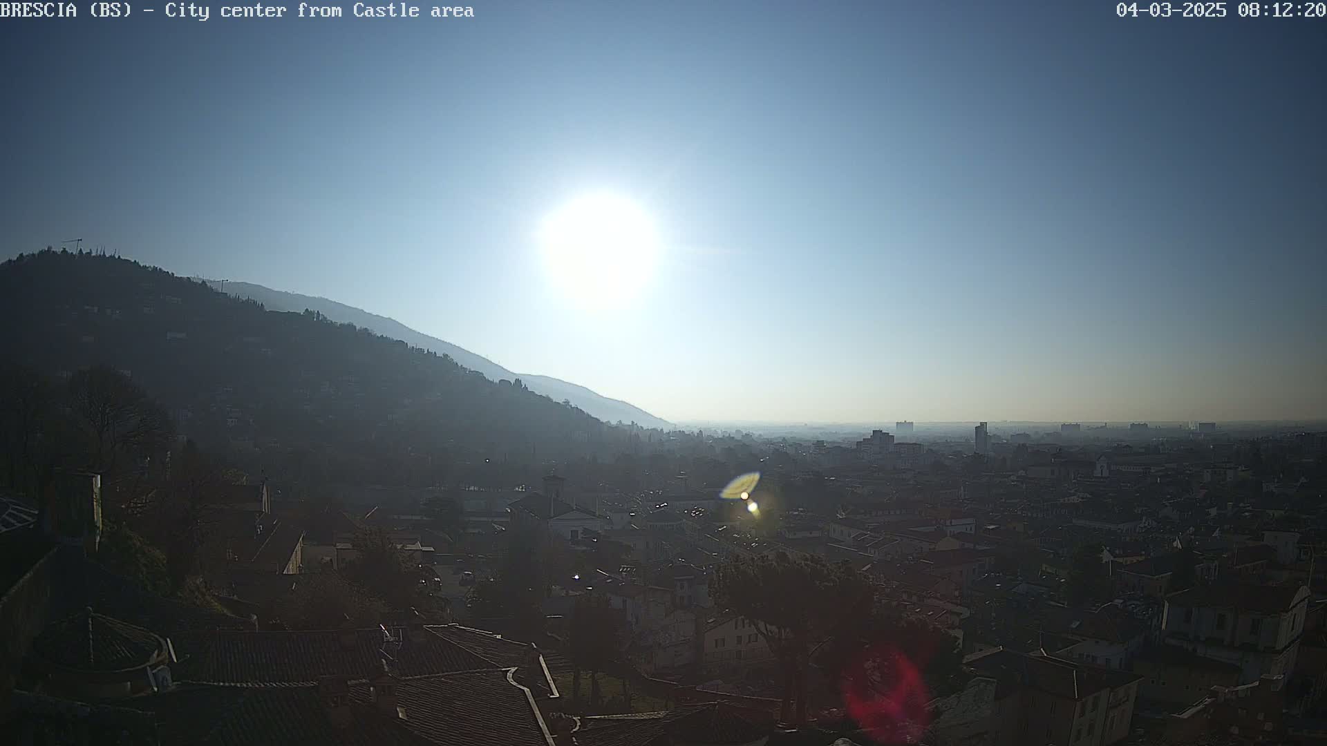 A hazy sun shines brightly over a city nestled at the base of a mountain, viewed from a high vantage point.