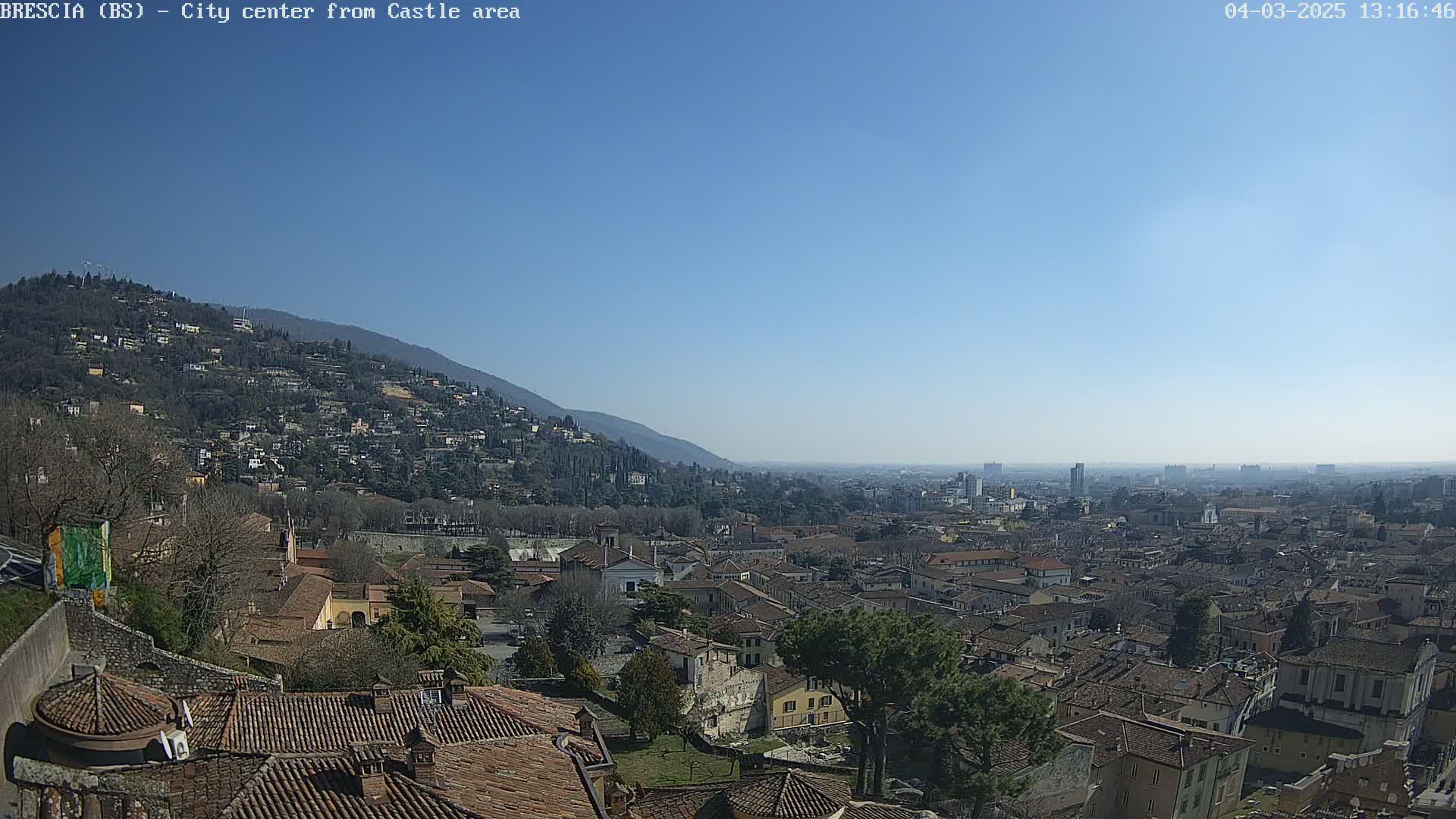 A sprawling city with red-tiled roofs nestles against a hillside under a clear, sunny sky.