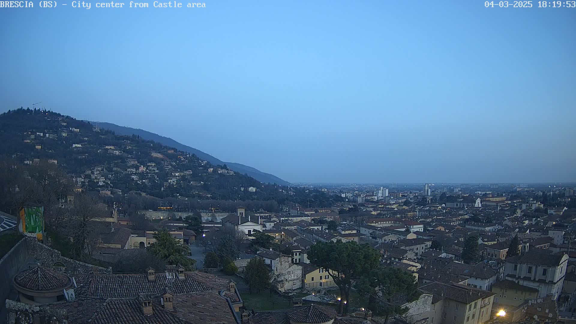 A sprawling city at dusk, nestled at the base of a hill dotted with houses, under a clear, twilight sky.