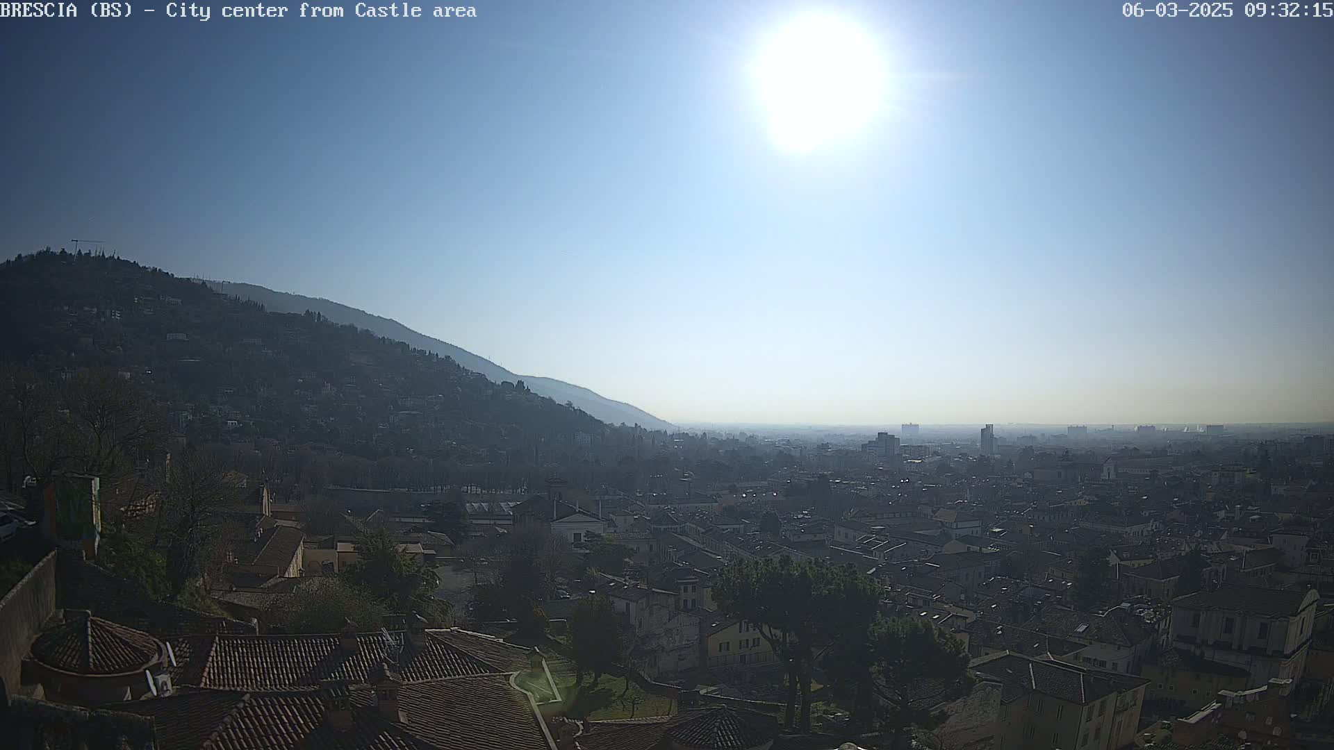 A sunny day reveals a sprawling city nestled between a hill and a hazy horizon, viewed from a higher vantage point.