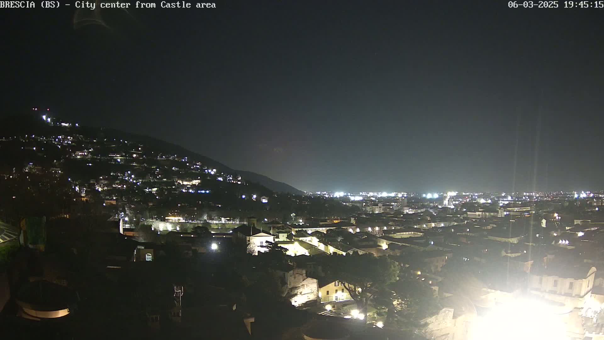 A nighttime, aerial view of a city nestled against a hill, illuminated by numerous lights under a dark sky.