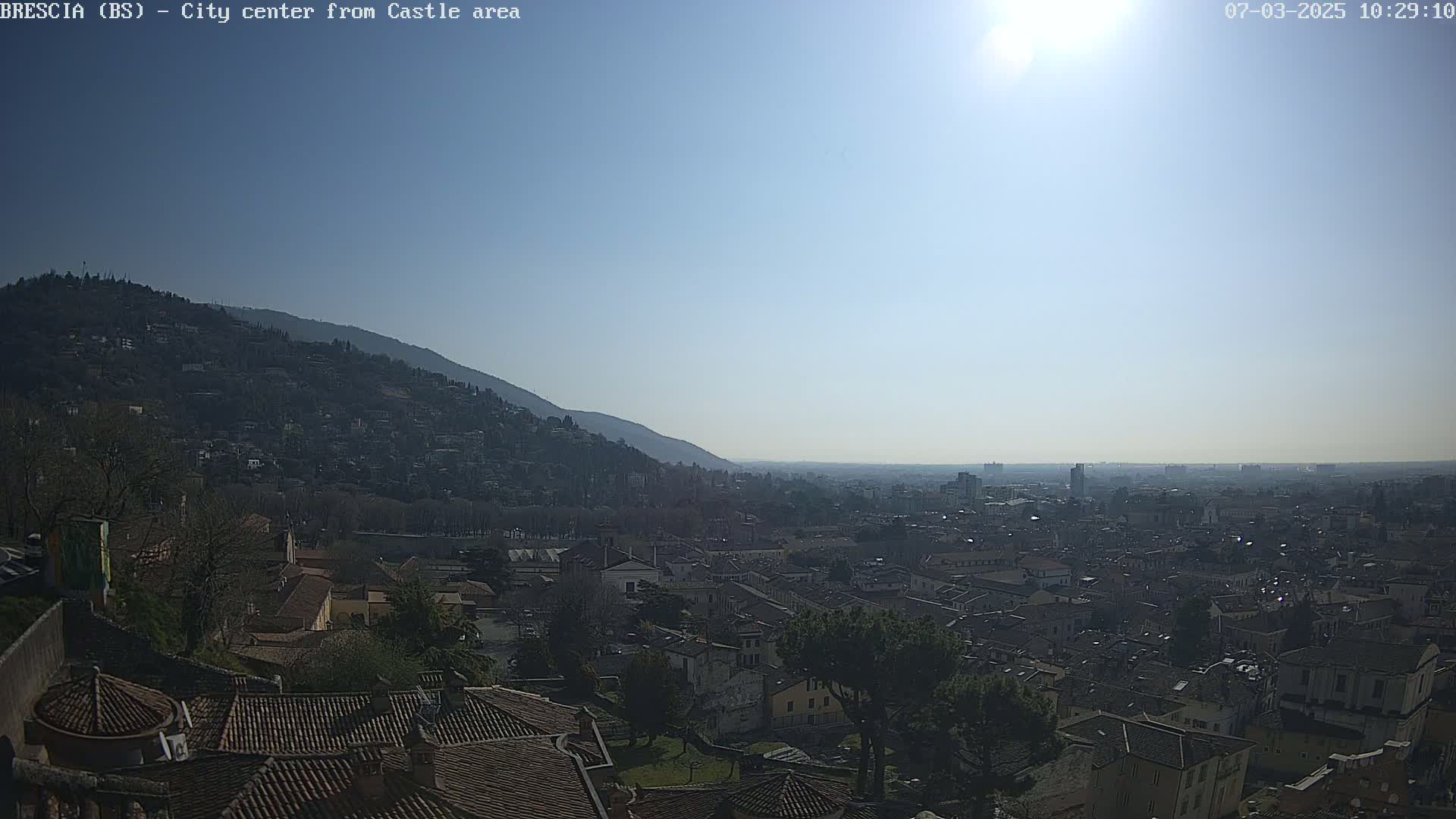 A sunny day reveals a sprawling city vista from a hilltop vantage point, showcasing a dense collection of buildings extending to a hazy horizon, with a mountain visible in the background.