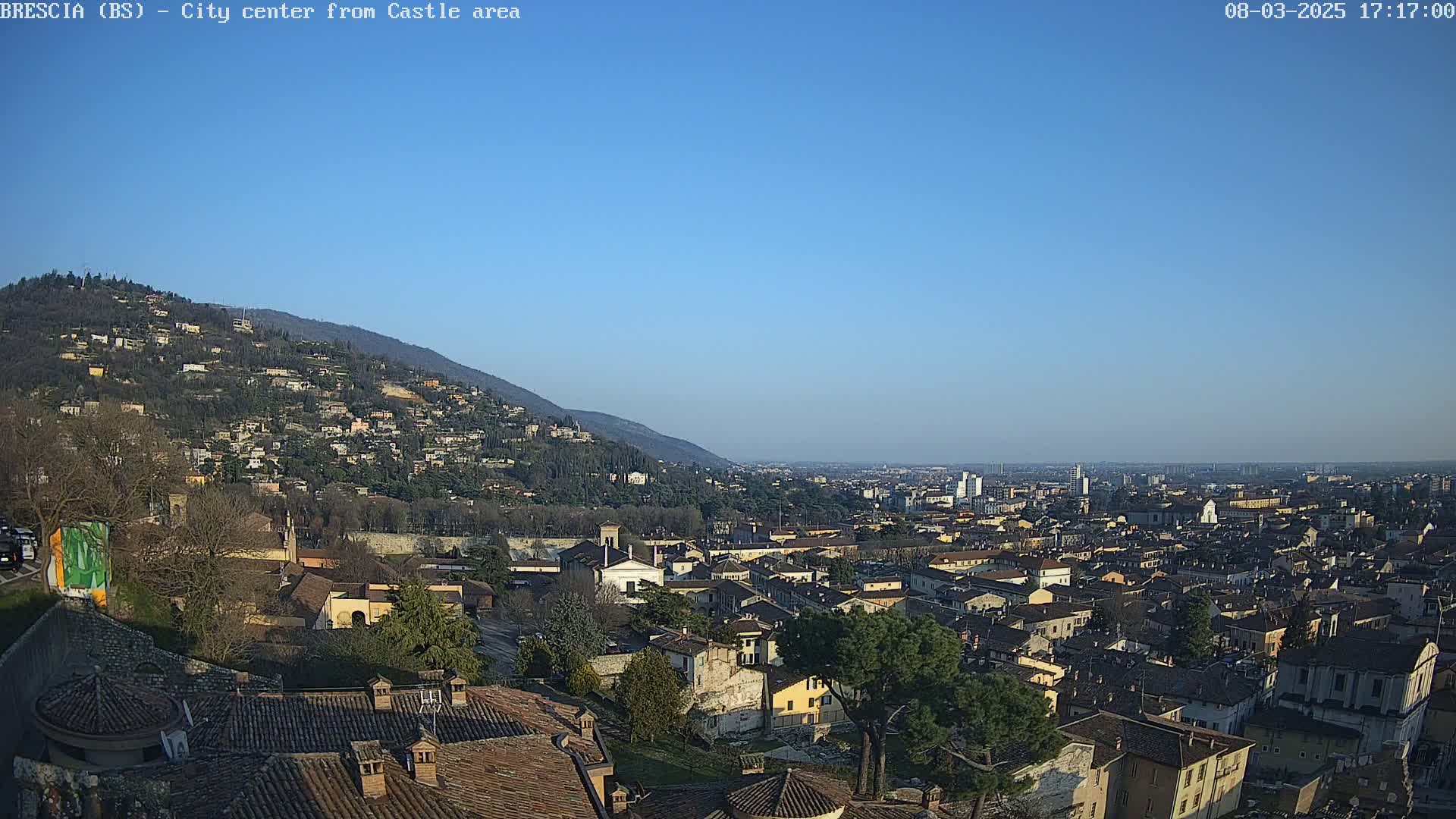 A sprawling city, nestled beneath a clear blue sky and a hillside dotted with buildings, is viewed from an elevated vantage point.