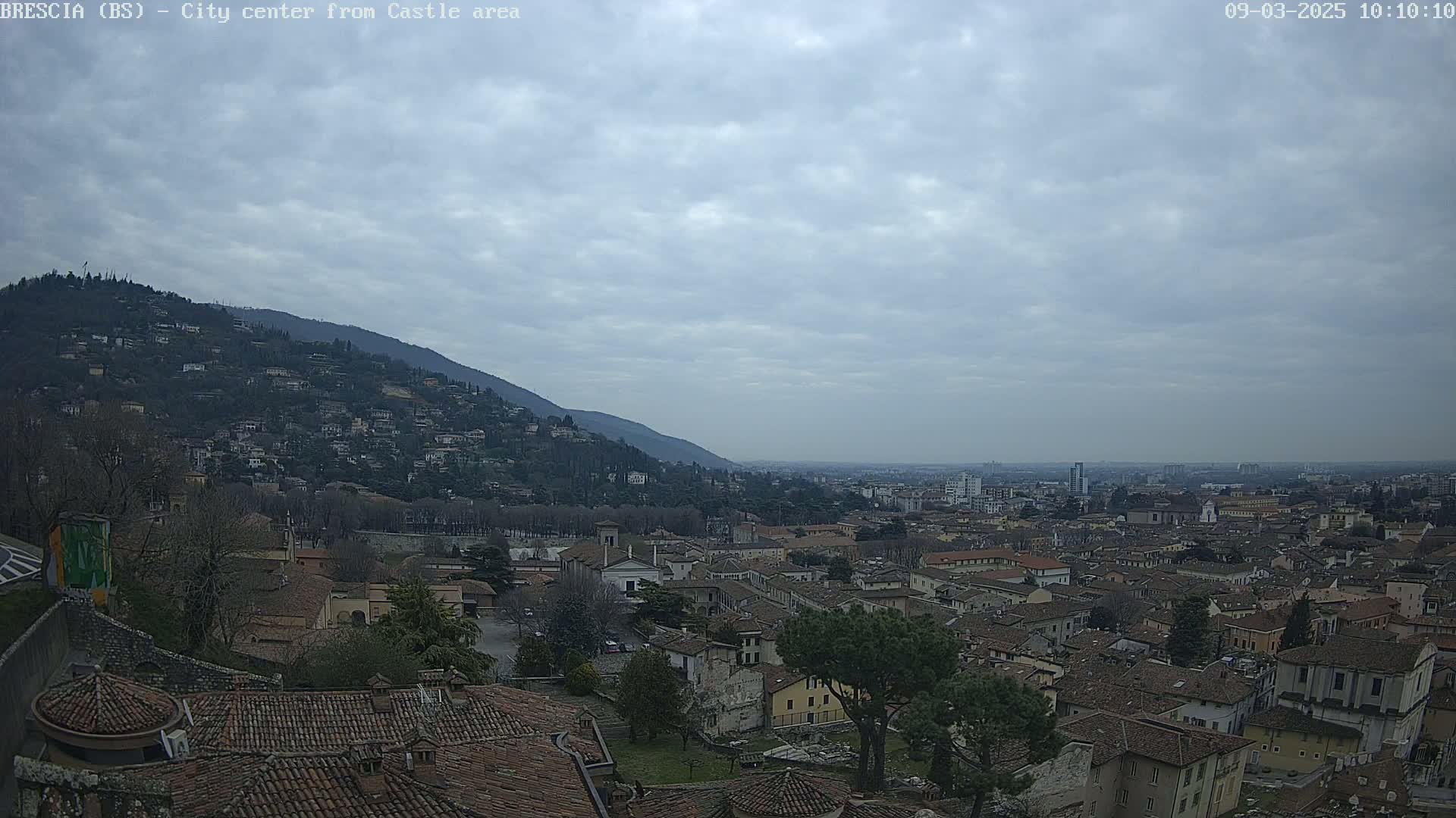 A sprawling city, nestled against a hillside under a cloudy sky, is viewed from an elevated vantage point.