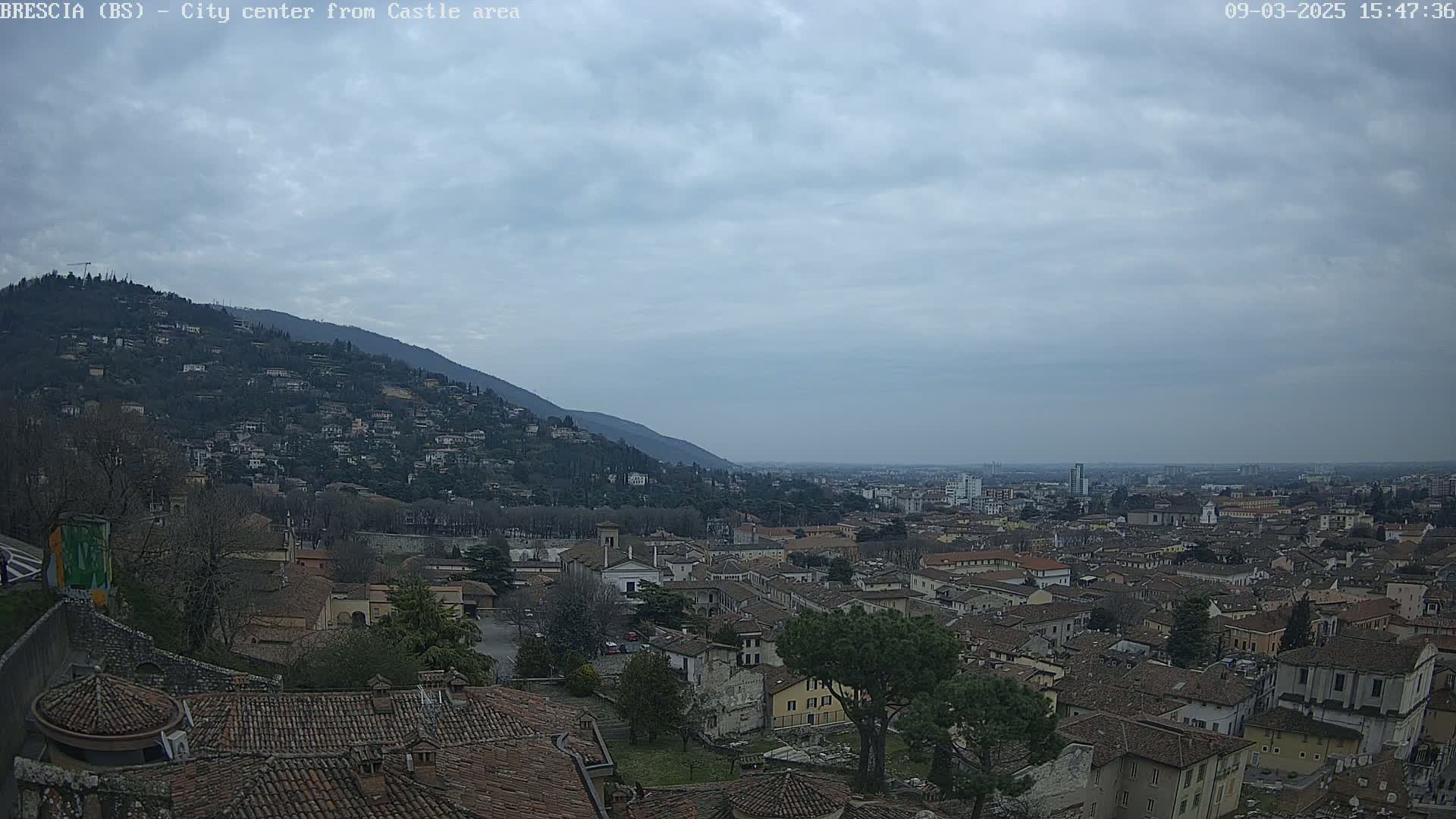A sprawling city with red-tiled roofs nestled against a hill under a cloudy sky.