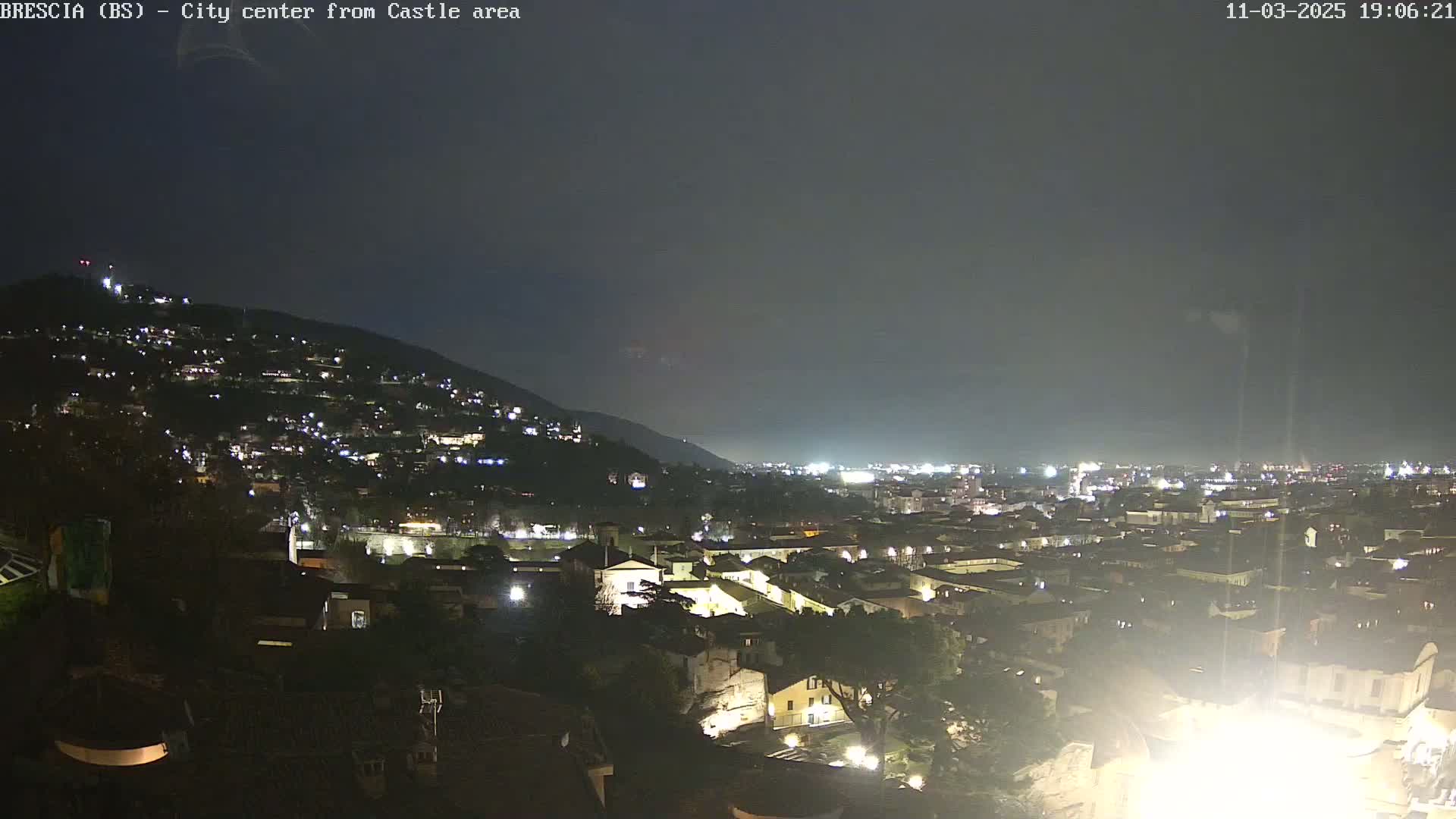 A nighttime, aerial view of a city nestled against a hill, under a dark, overcast sky.