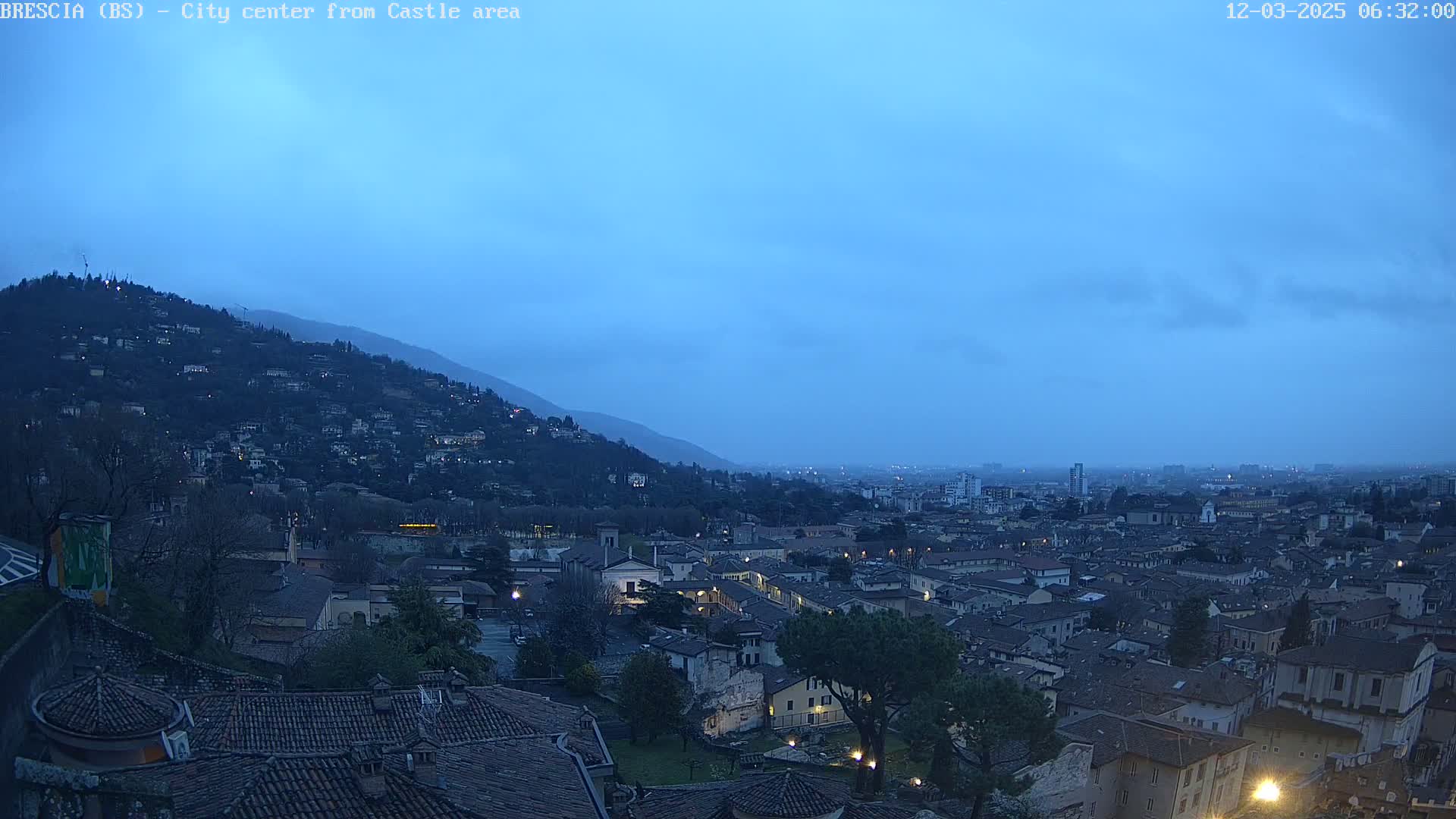 A hilltop view of a city at twilight under a mostly clear, blue-ish sky, showing many buildings with lights beginning to appear.