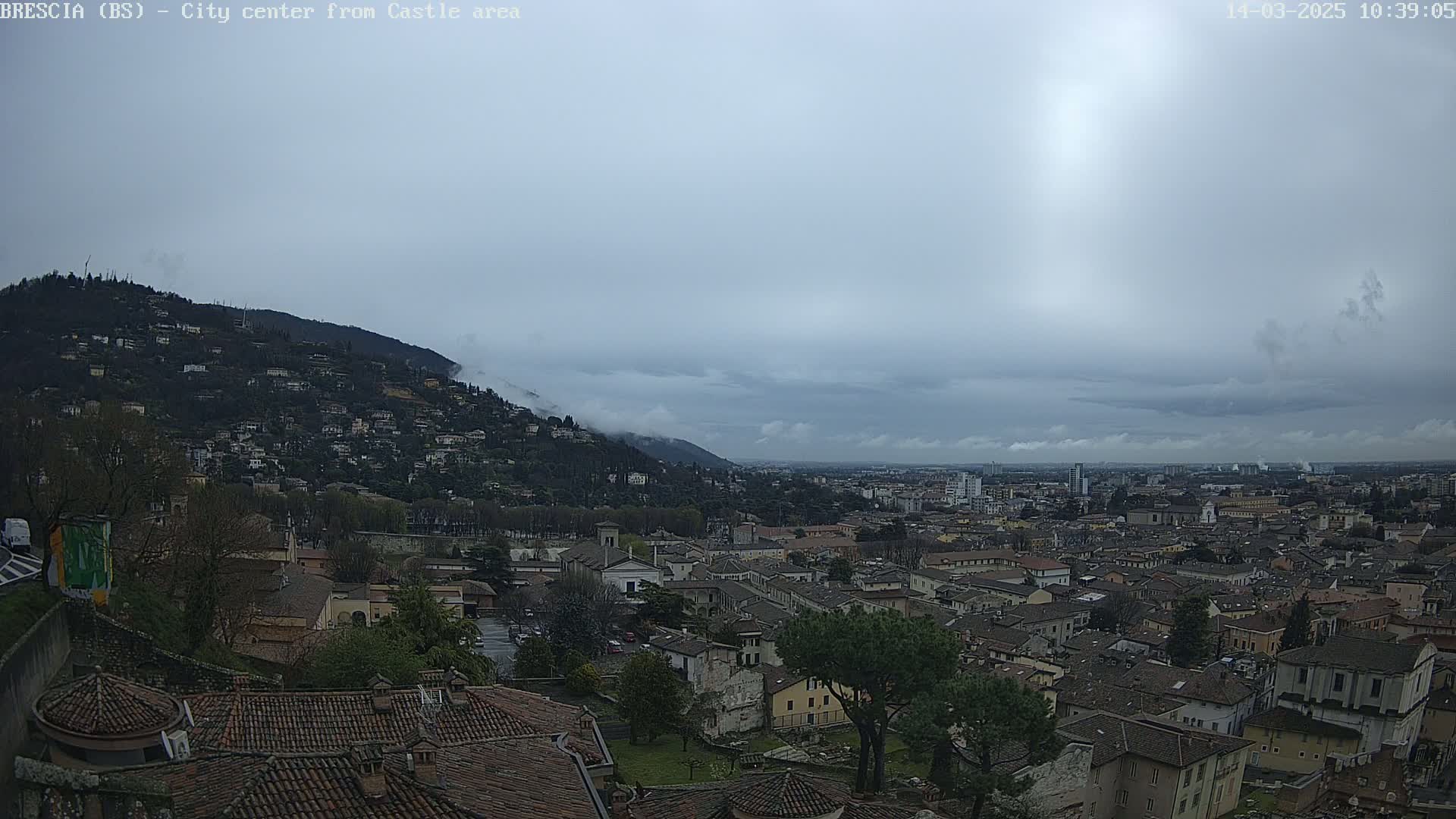 A sprawling city with low-lying buildings stretches out beneath a cloudy, overcast sky, nestled against a hill shrouded in fog.