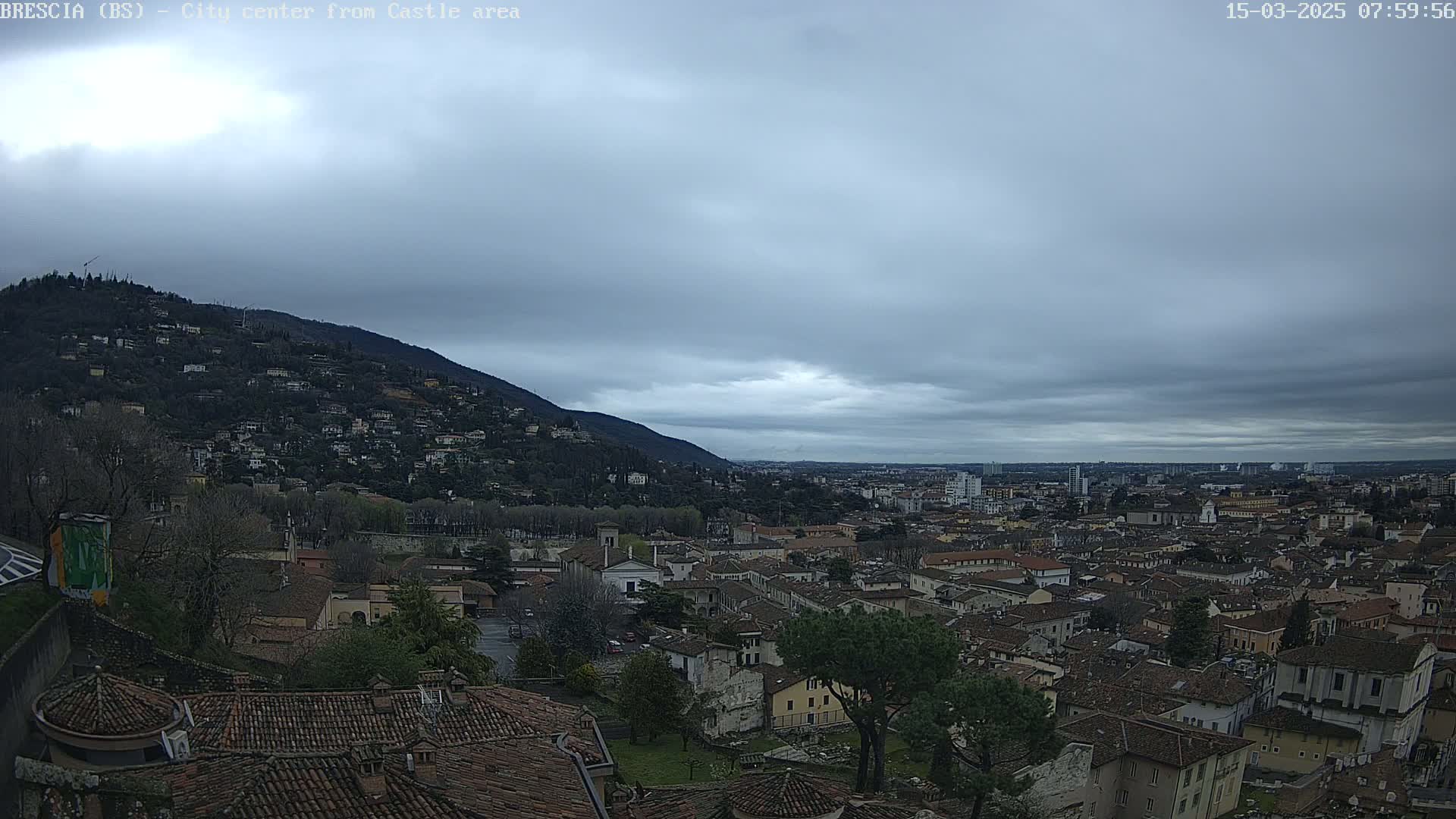 A sprawling city with terracotta rooftops nestled beneath a cloudy, overcast sky and a hill dotted with buildings in the background.