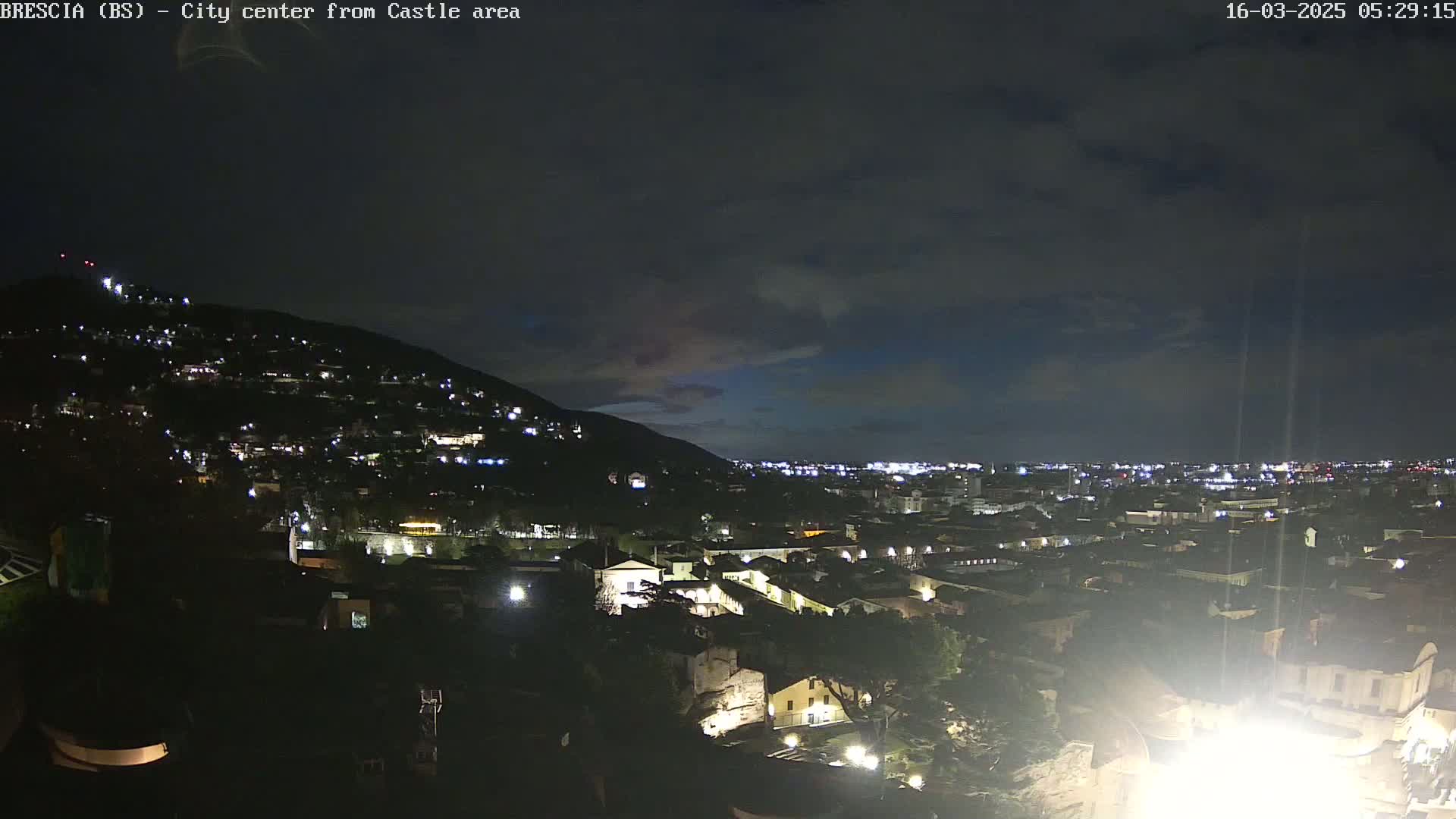 A nighttime view of a city nestled against a hill, under a mostly cloudy sky.