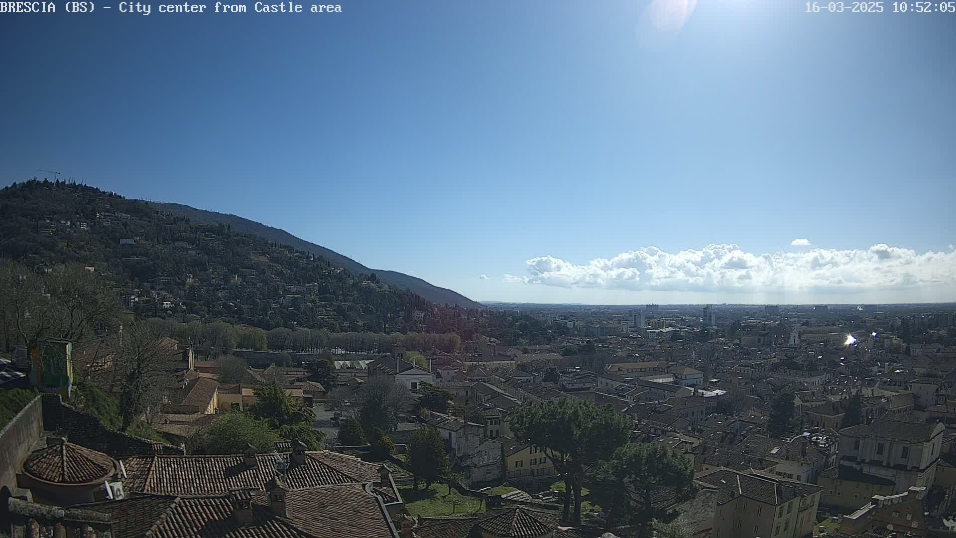 A sunny day reveals a sprawling cityscape nestled beneath a hill, with puffy white clouds scattered across a mostly clear blue sky.