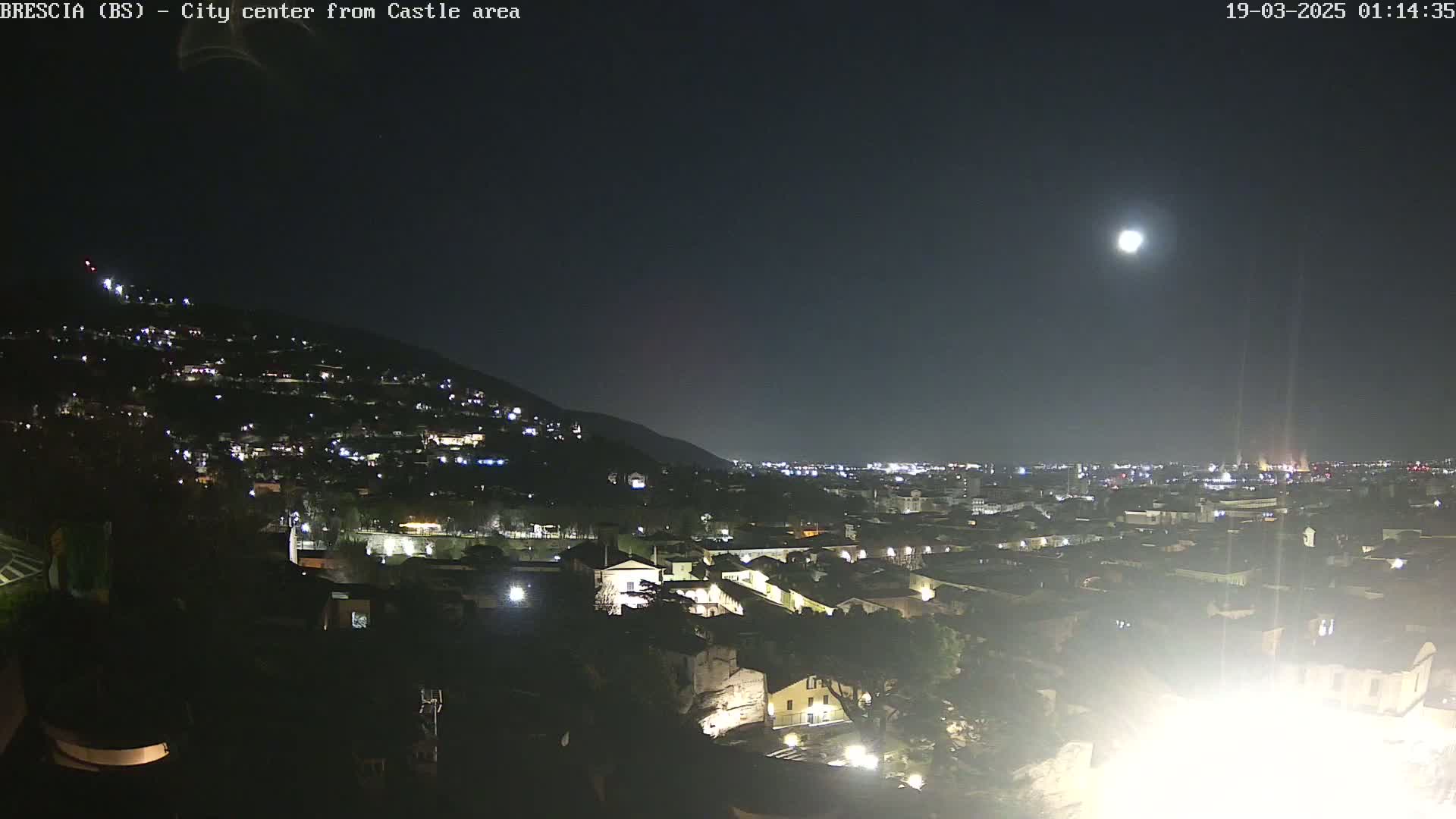A nighttime view of a city nestled against a hill, illuminated by the lights of buildings and a bright moon in the clear night sky.