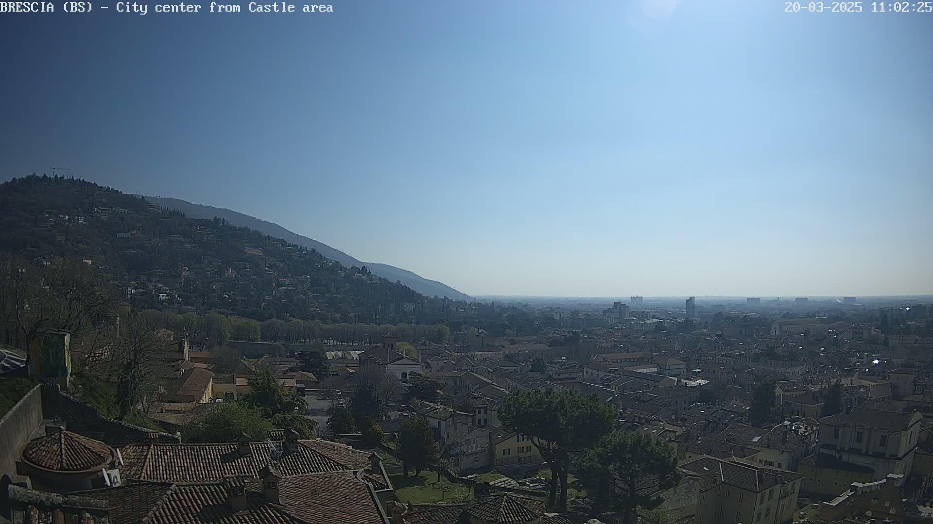 A sunny day reveals a sprawling city nestled at the foot of a mountain, viewed from a higher vantage point with terracotta rooftops dominating the foreground.