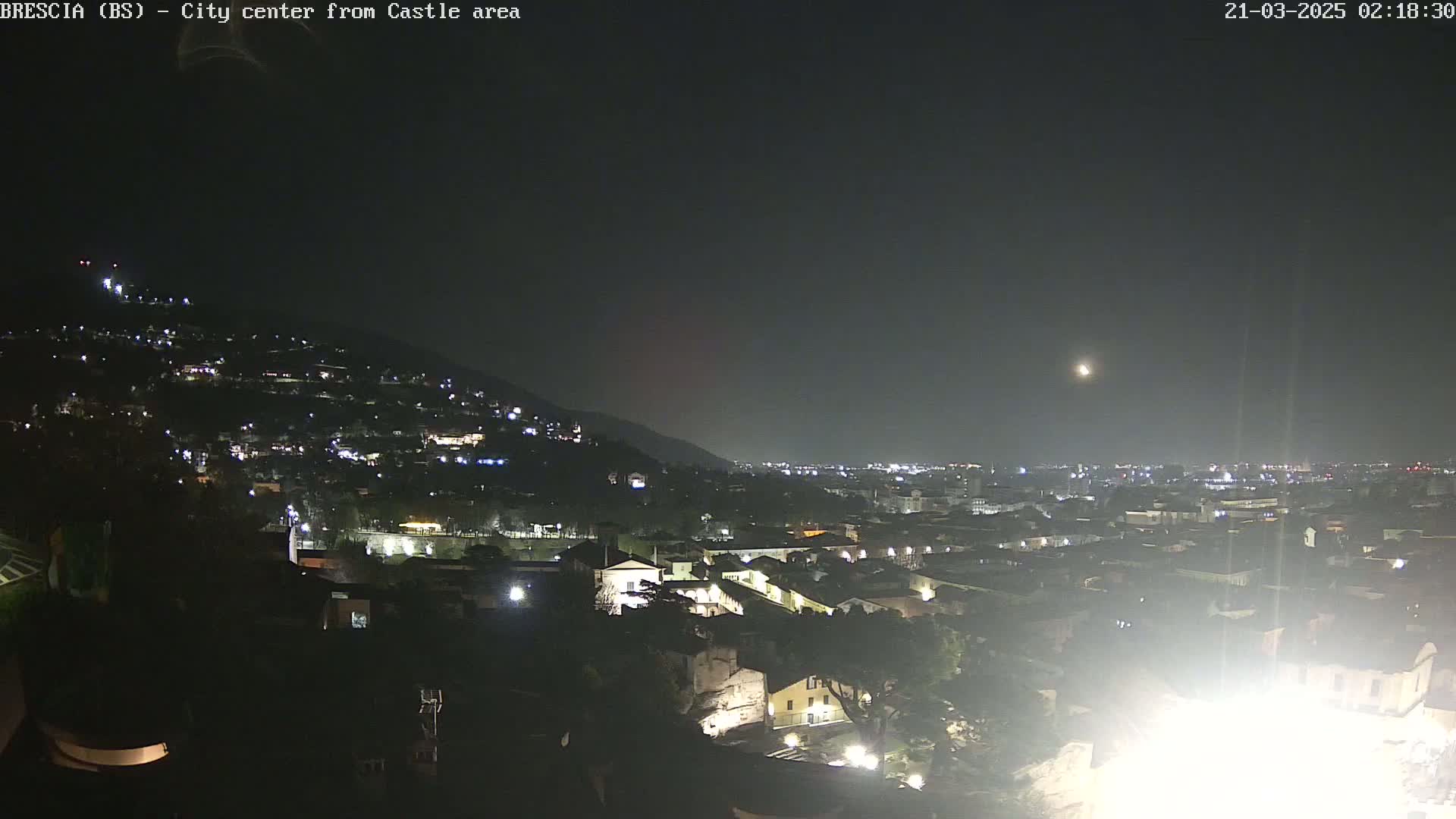 A nighttime view of a city nestled against a hillside, illuminated by scattered lights under a clear, moonlit sky.