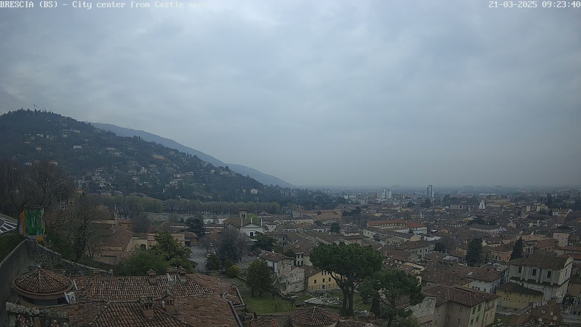 A hazy, overcast day reveals a sprawling city nestled against a mountainside, viewed from an elevated vantage point, showing a landscape of terracotta rooftops and green spaces.