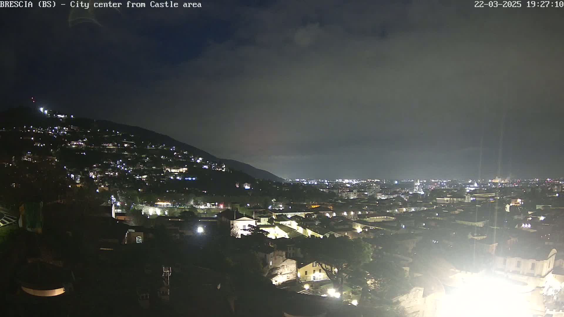 A nighttime view of a city nestled in a valley beneath a hill, under a mostly cloudy sky.