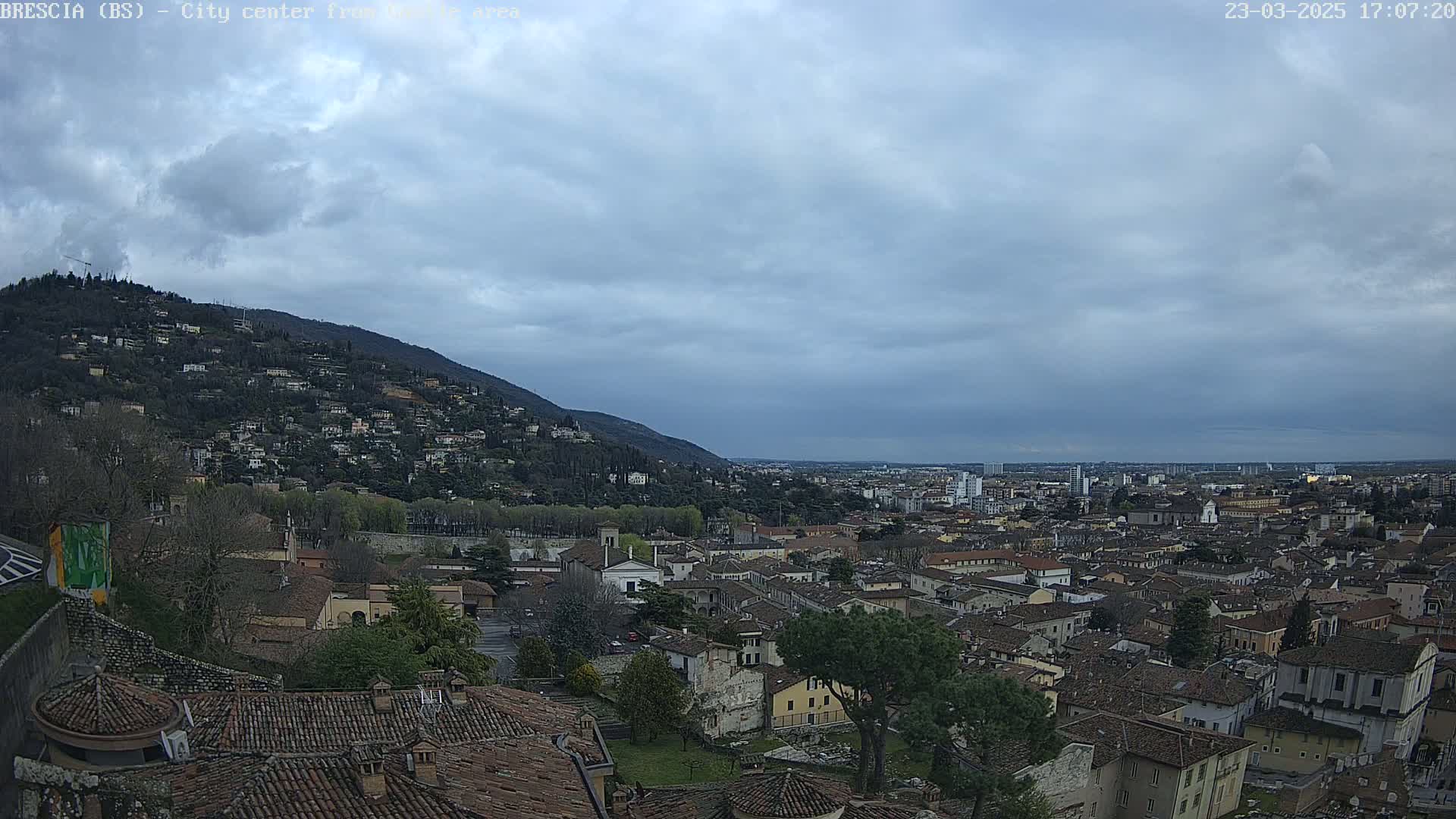 A sprawling city nestled beneath a hill, viewed from an elevated vantage point under a mostly cloudy sky.