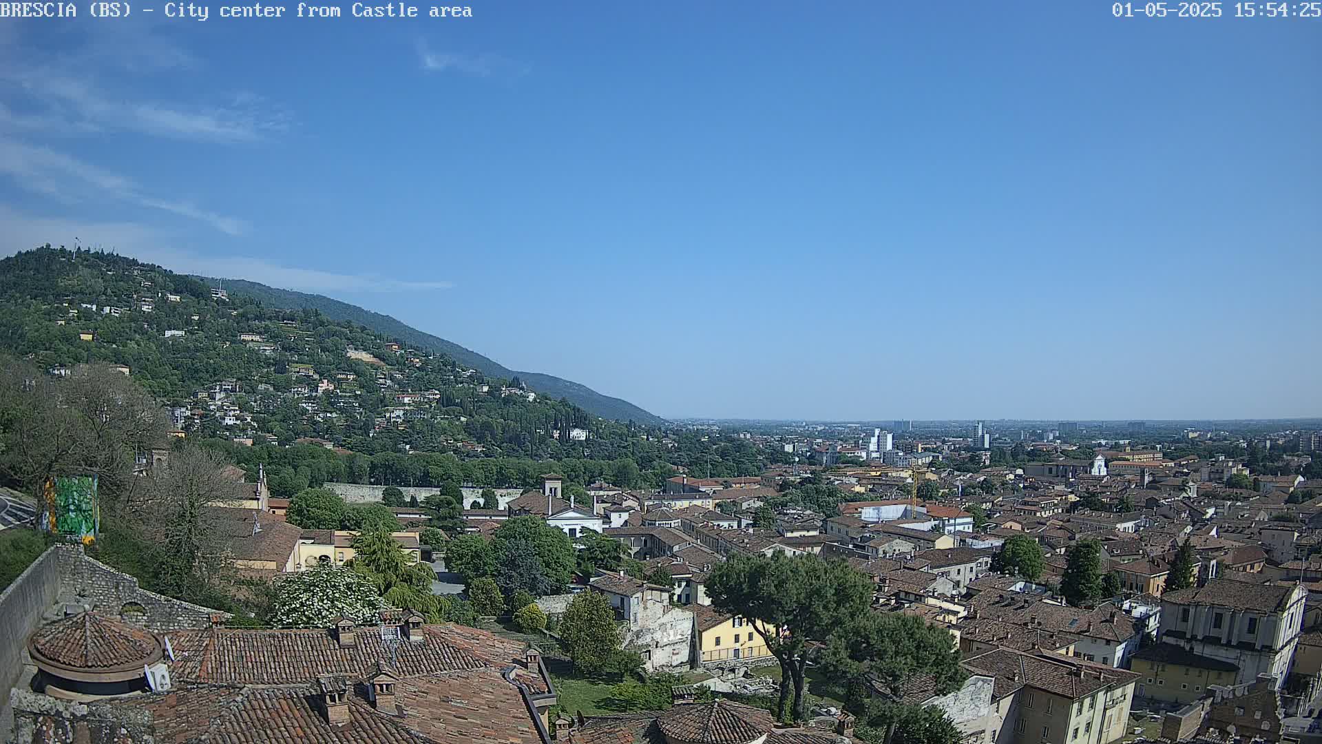 A sprawling city with red-tiled roofs nestles beneath a verdant hillside under a clear blue sky.