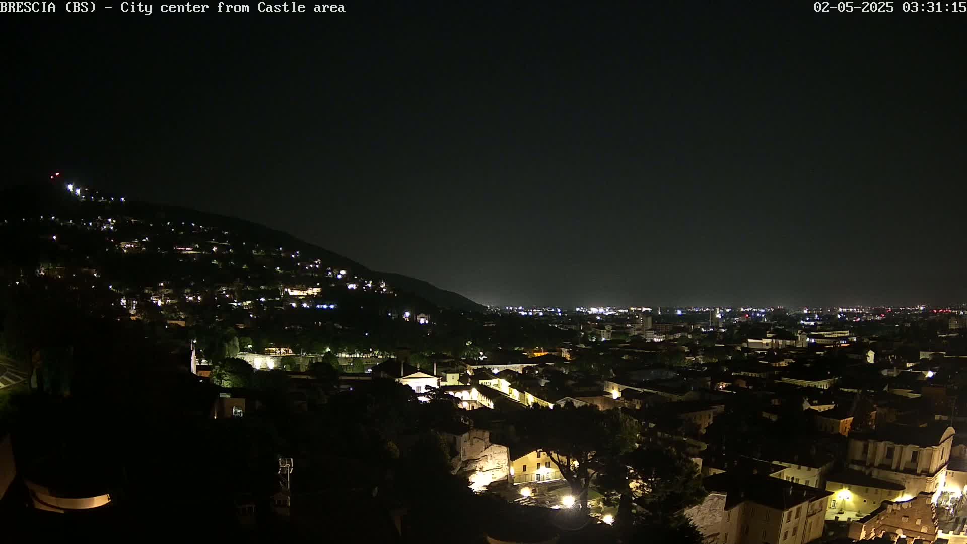 A nighttime, aerial view shows a city nestled against a hillside, illuminated by scattered lights under a dark sky.
