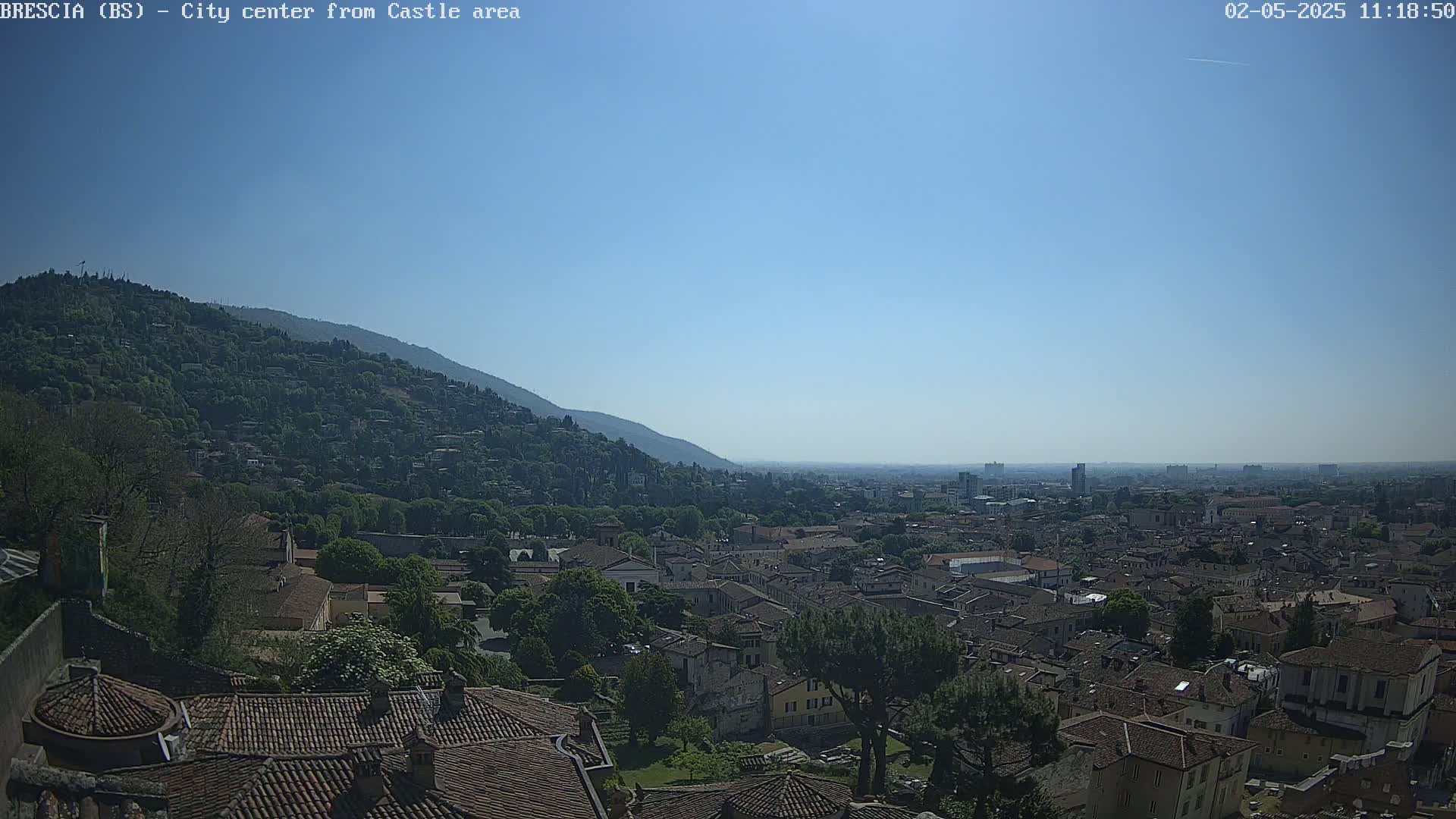 A sprawling city with terracotta rooftops nestled against a verdant hillside under a clear, bright blue sky.