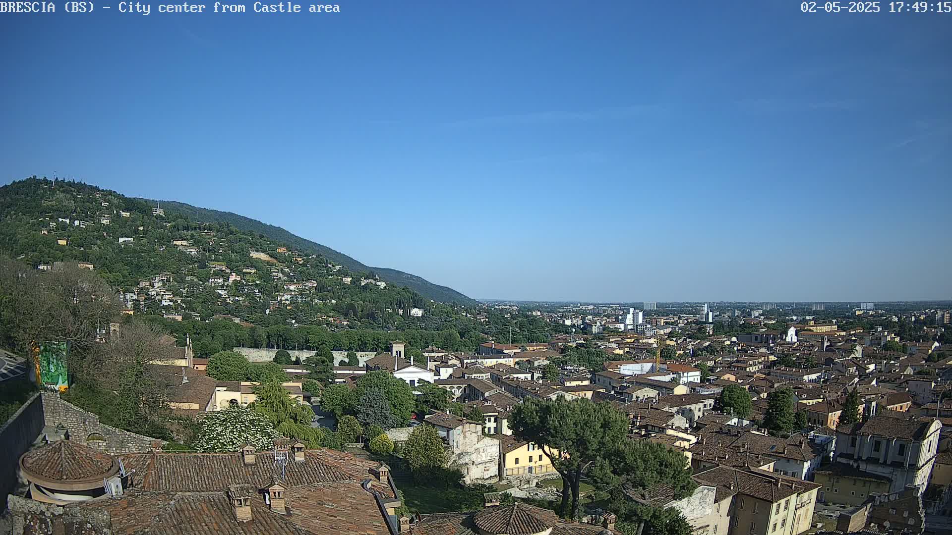 A sprawling city landscape, nestled against a verdant hillside under a clear blue sky, is viewed from an elevated position.