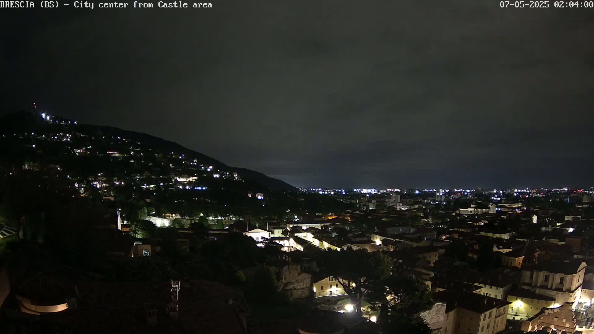 A nighttime view of a city nestled in a valley, illuminated by numerous lights, under a dark, cloudy sky.