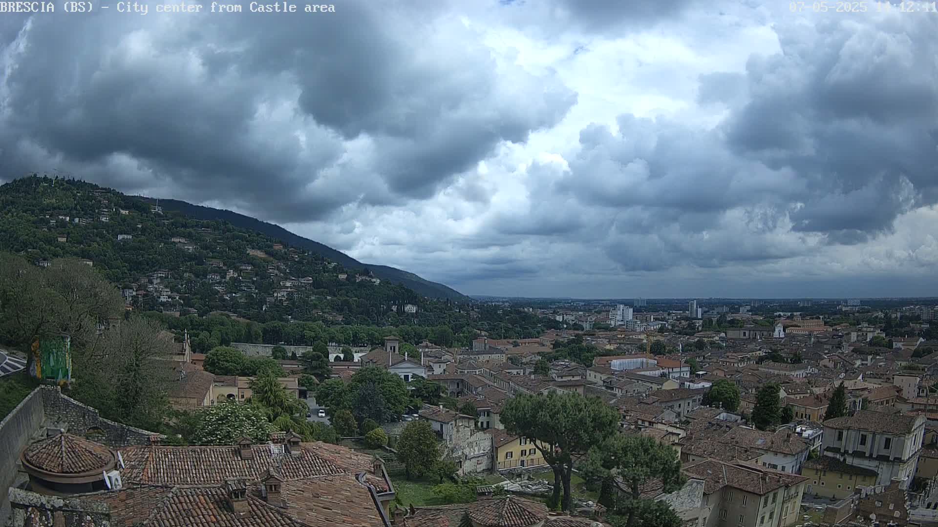 A sprawling city with red-tiled roofs nestles beneath a cloudy sky, backed by a verdant hill.