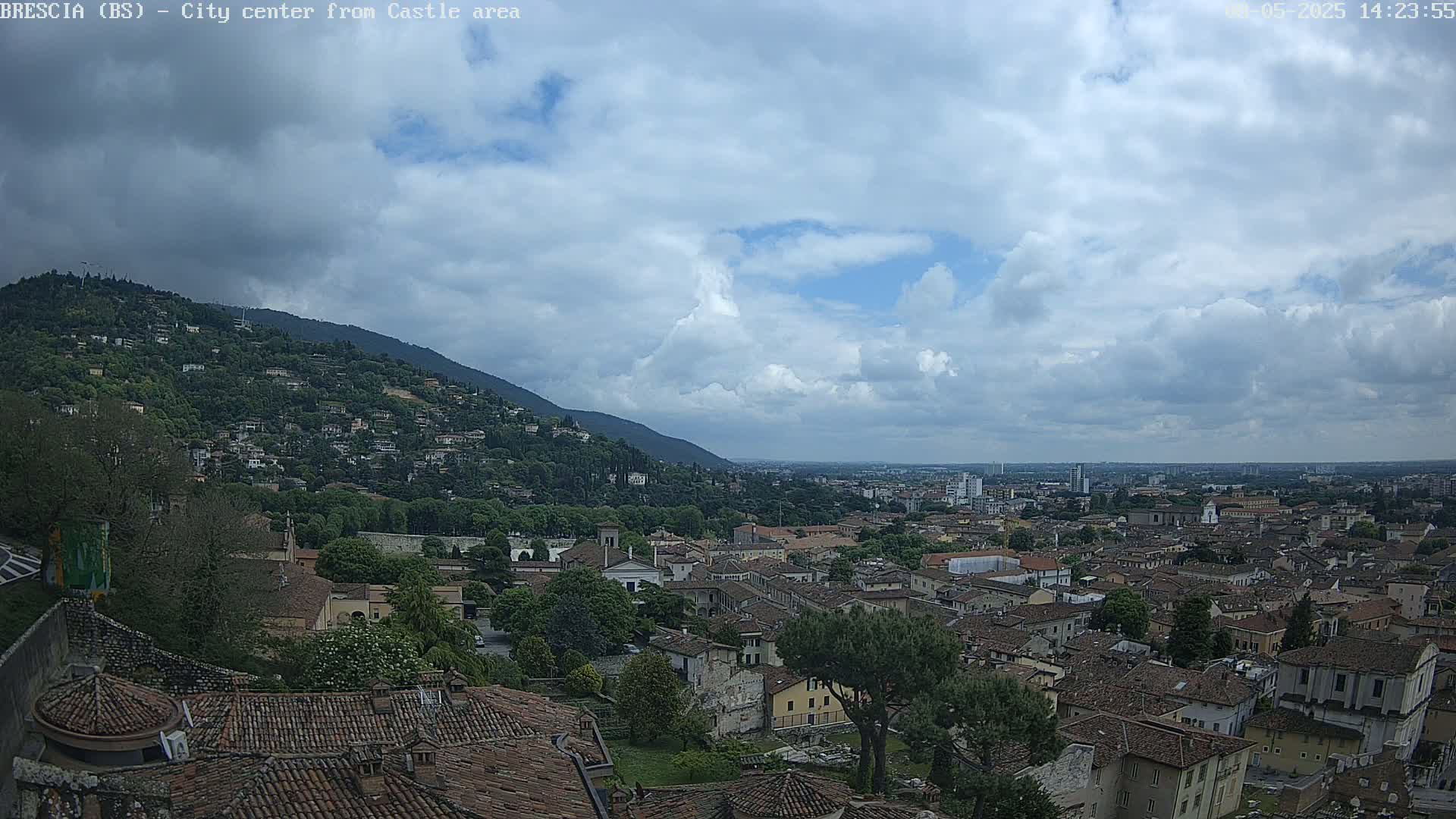 A sprawling city with many brown-roofed buildings nestles in a valley below a hill covered in houses and trees, under a partly cloudy sky.