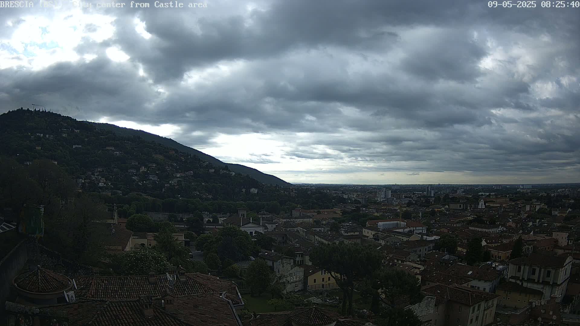 A sprawling city with red-tiled roofs nestles beneath a cloudy sky, with a hill covered in dense vegetation in the background.