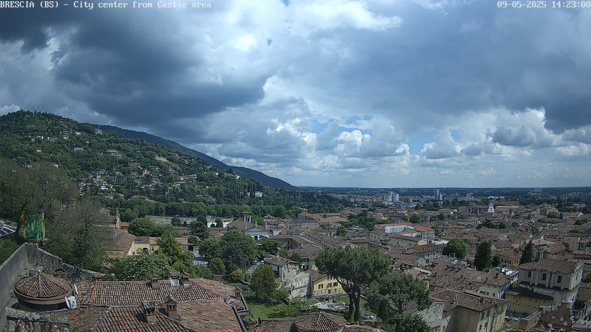 A sprawling city with terracotta rooftops nestled against a green hillside under a partly cloudy sky.