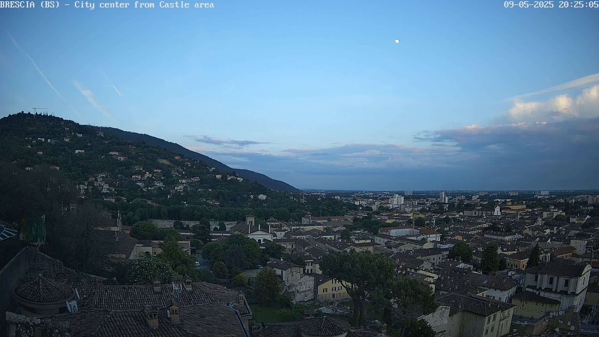 A moonlit cityscape at dusk, nestled beneath a hill covered in vegetation, with a partly cloudy sky.