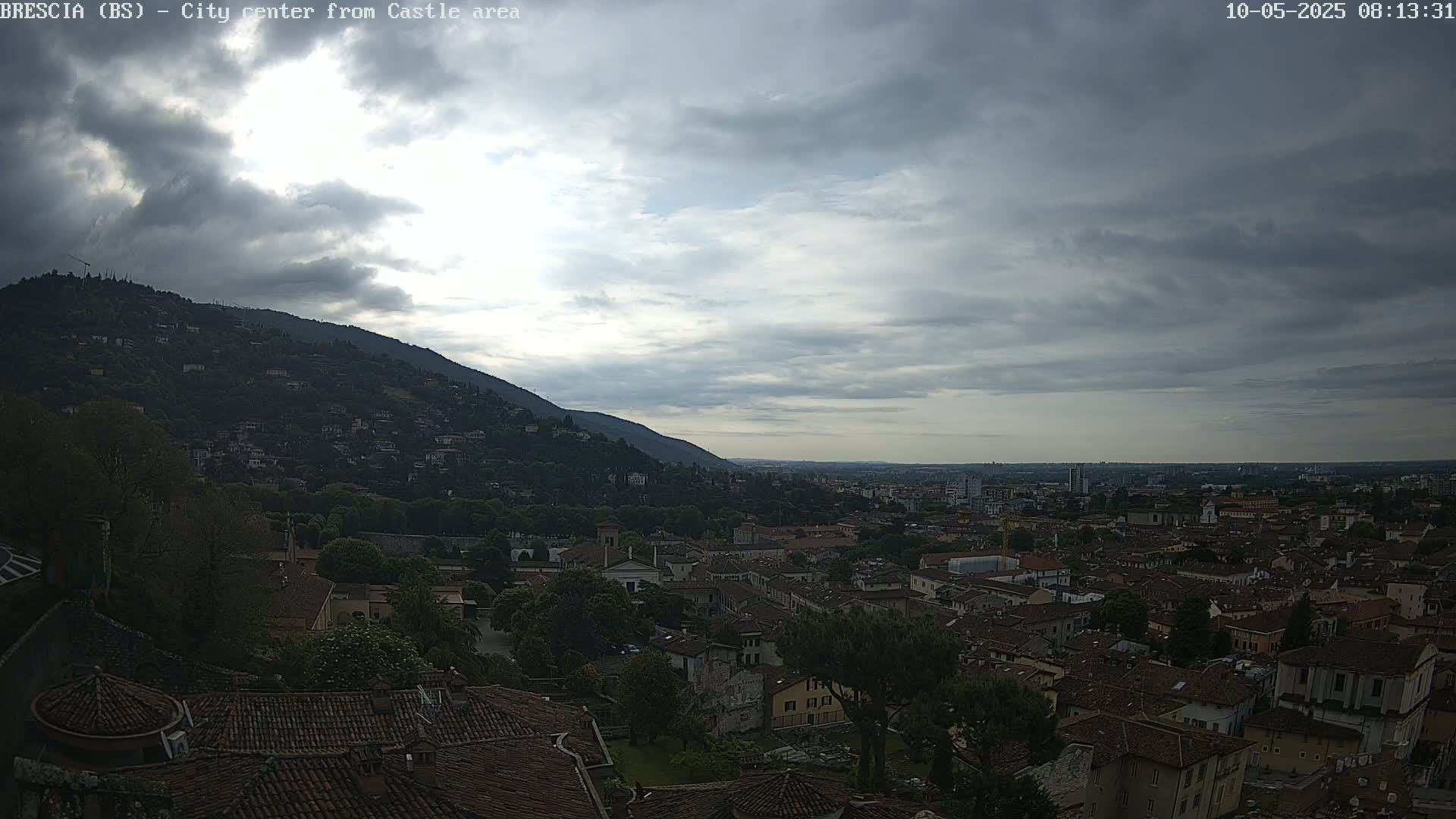 A sprawling city with red-tiled roofs nestled at the foot of a verdant hill under a mostly cloudy sky.