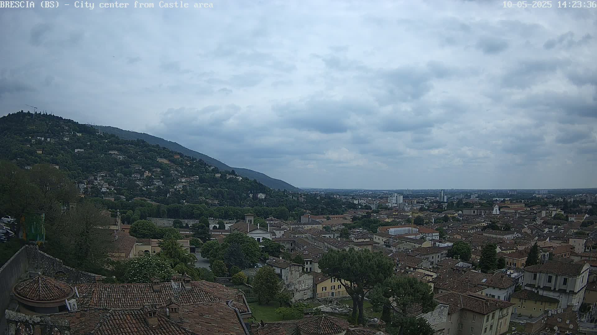 A sprawling city with terracotta rooftops nestled against a green hill under a mostly cloudy sky.