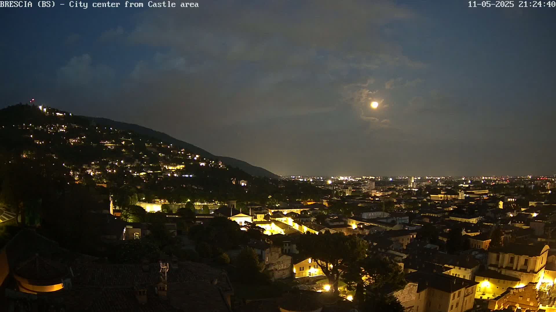 A nighttime view of a city nestled against a hillside, illuminated by lights and a bright moon in a partly cloudy sky.