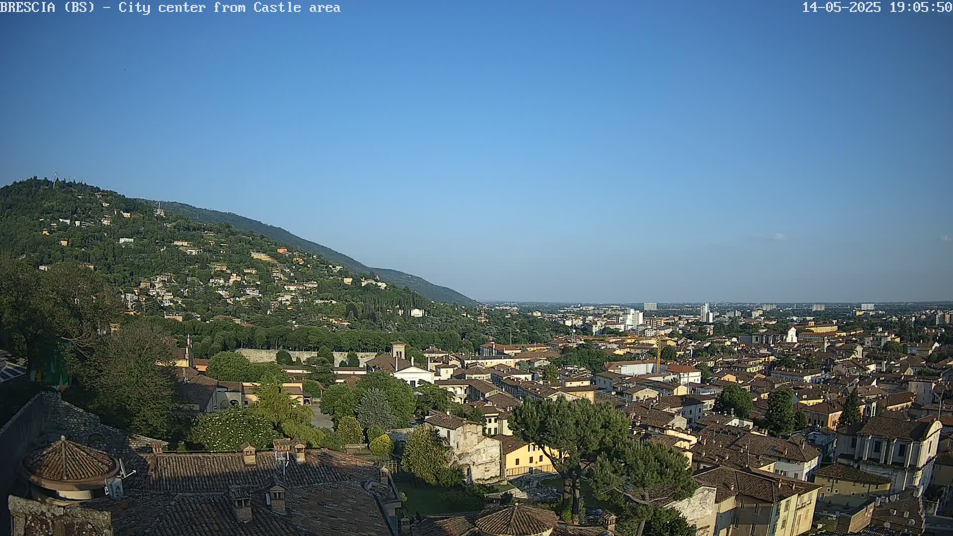 A sprawling city, nestled against a verdant hillside under a clear blue sky, is viewed from an elevated vantage point.