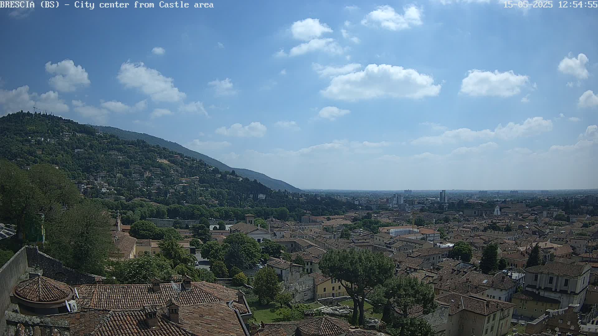 A partly cloudy sky overlooks a sprawling city of terracotta rooftops nestled against a verdant hillside.