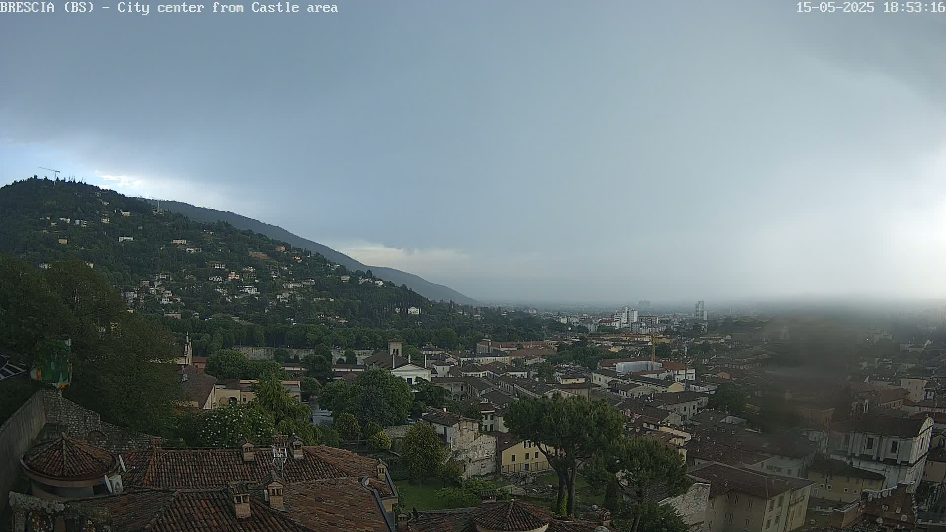 A hazy, overcast view shows a city nestled in a valley between a hill covered in buildings and a larger mountain in the background.