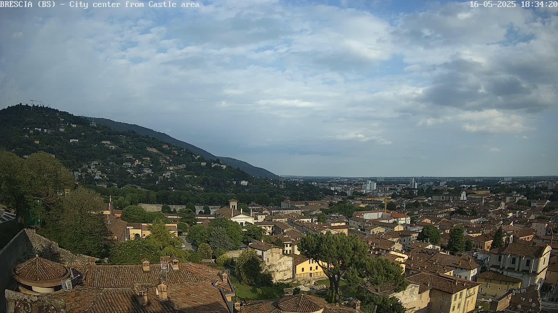 A sprawling city with terracotta rooftops nestles against a verdant hillside under a partly cloudy sky.