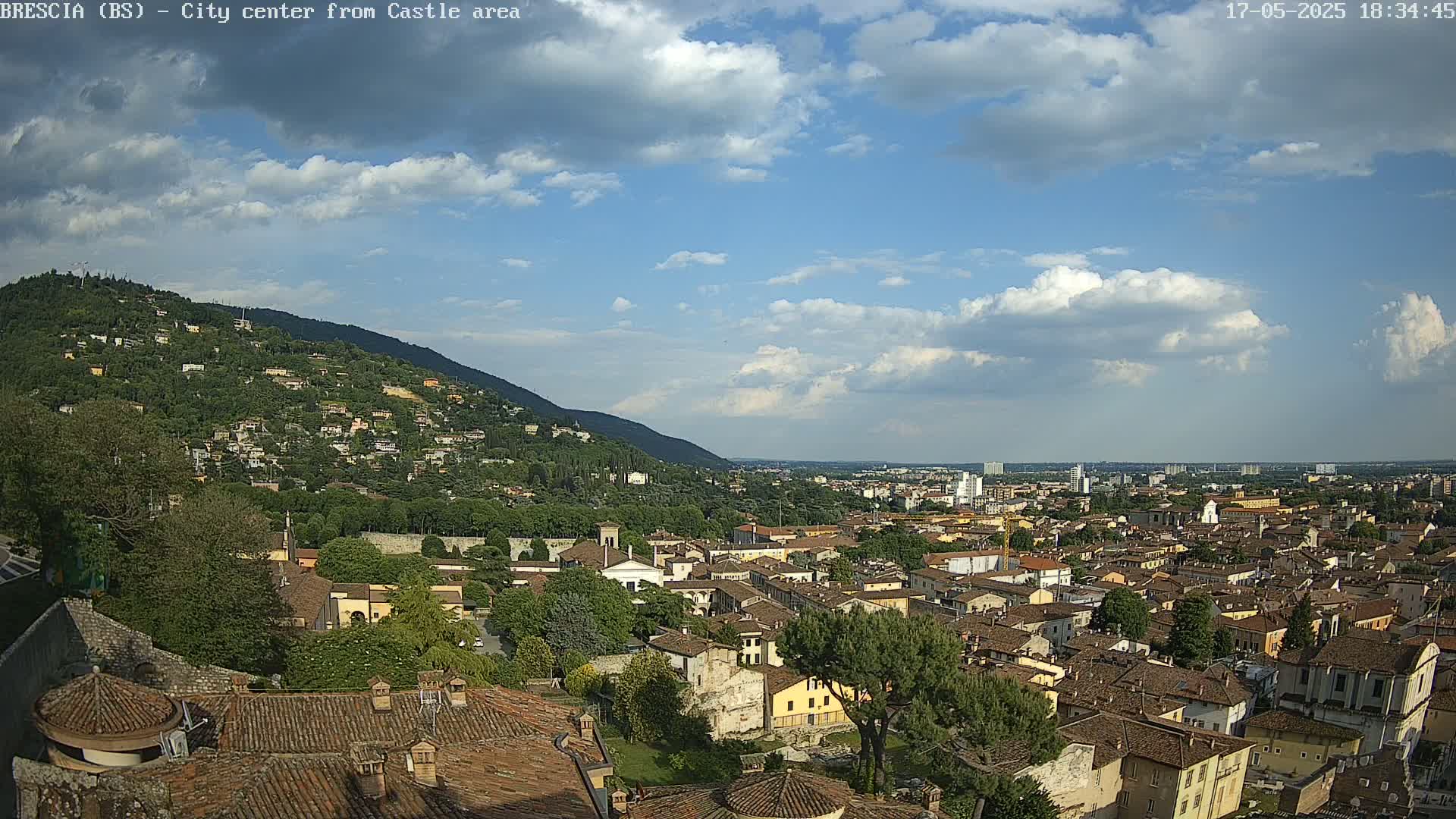 A sprawling city with numerous terracotta-roofed buildings nestles beneath a hill, all under a partly cloudy sky.