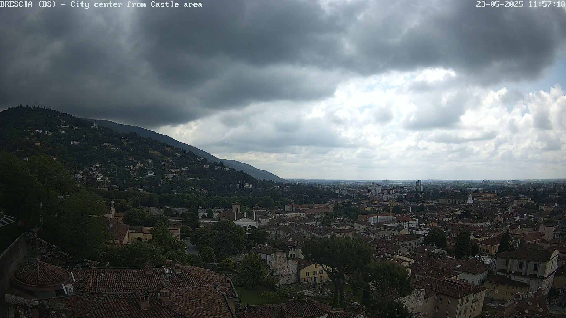 A city sprawls beneath a cloudy sky, with a hill covered in buildings and trees in the foreground.